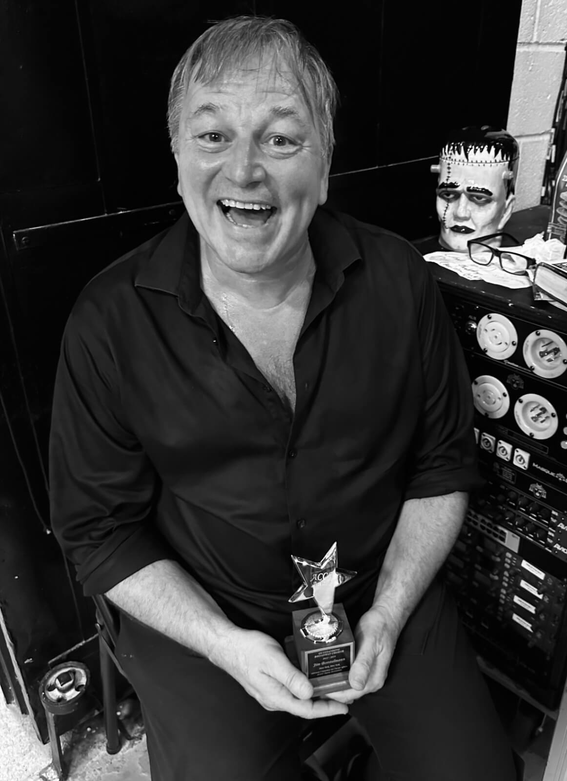 Black and white photo of Jim Borstelmann seated backstage at New York, New york after winning the ACCA award for Outstanding Ensemble on Broadway. He holds award in his lap and sports a big open mouth smile.