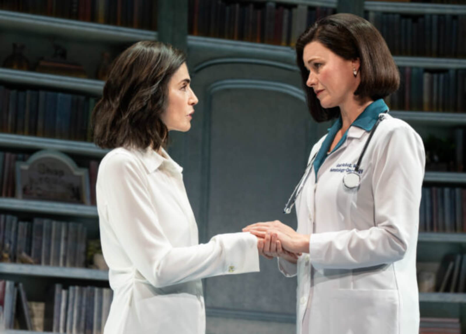 Julianna Margulies (looking concerned) has her hand being held by Kate MacCluggage (dressed as a doctor with a stethoscope). 