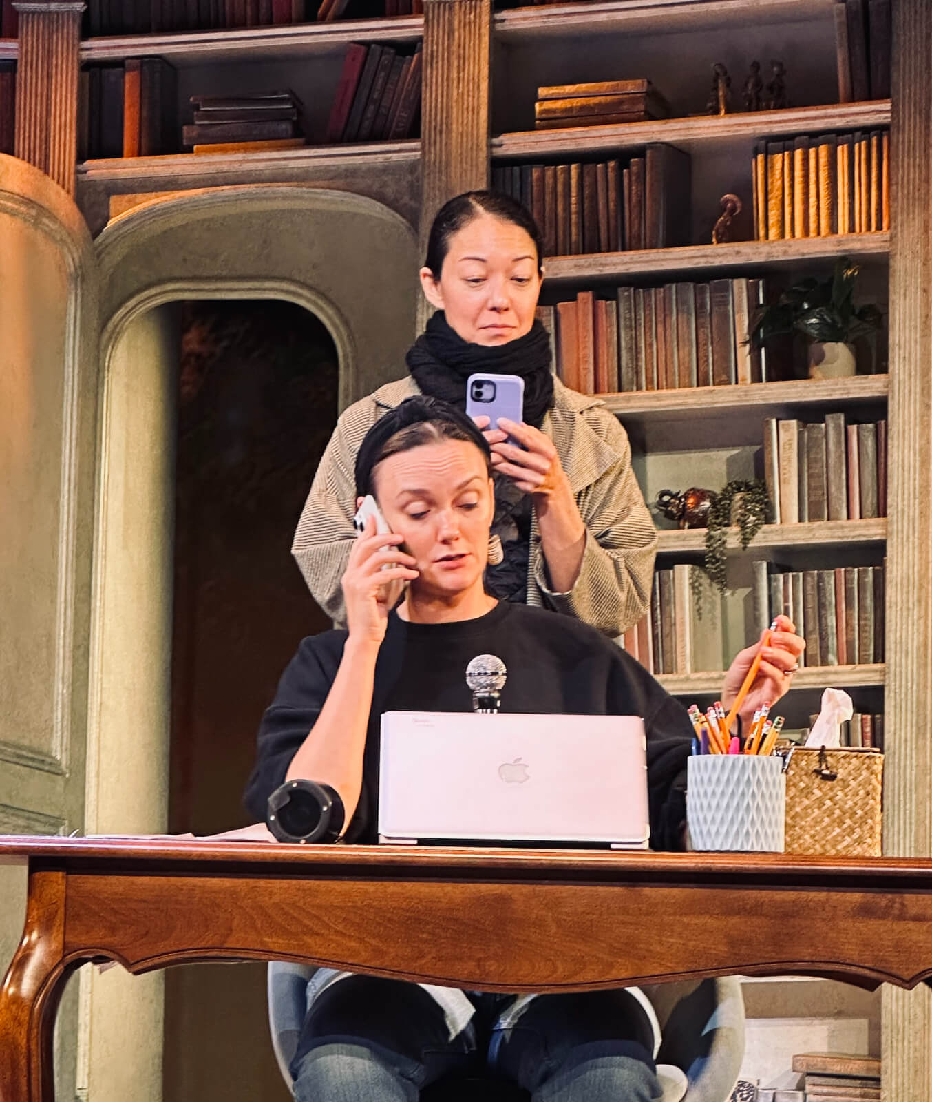 Associate Director Leah Hofmann and Assistant Director Naomi Kakuk in rehearsal for Left on Tenth. They are on the set. Leah is seated a desk with a MacBook while speaking into an iPhone on her right ear. Naomi is standing behind her texting on an iPhone.