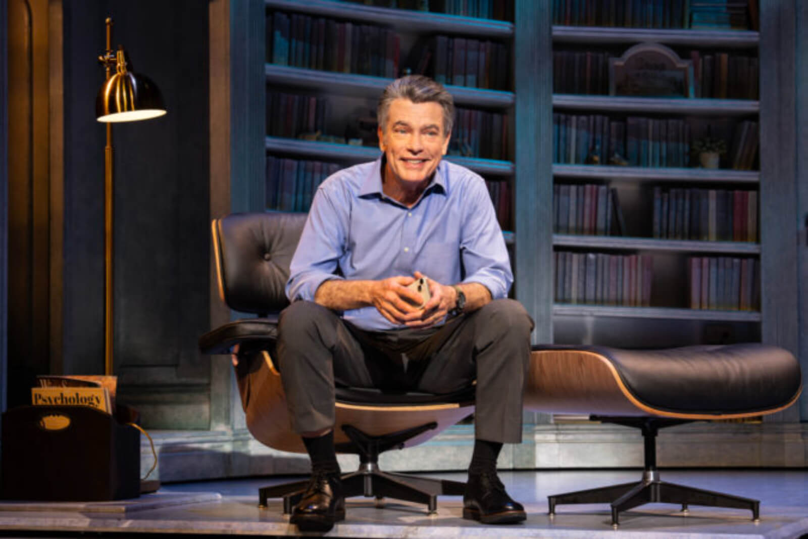 Peter Gallagher is seated in a black leather chair downstage with iPhone in his hands and psychology magazines in a rack next to him. 