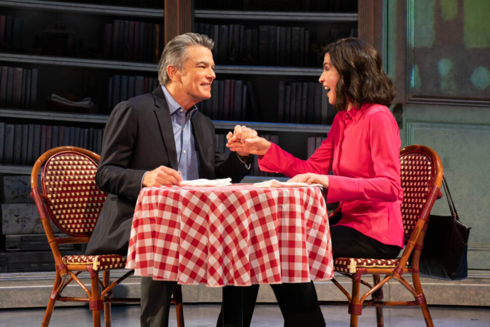Peter Gallagher and Julianna Margulies seated on a red and white checkered tablecloth holding hands. 
