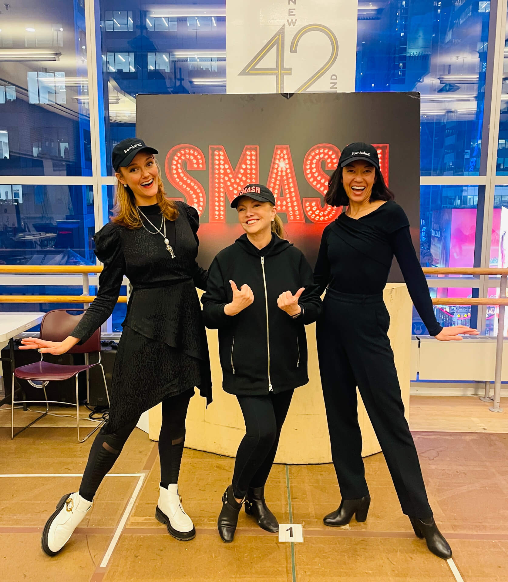 Color photo of SMASH poster behind Associate Director Leah Hofmann, Director Susan Stroman, and Assistant Director-Choreographer Naomi Kakuk. The girls are posed around Stro who is pointing at them with two thumbs up.
