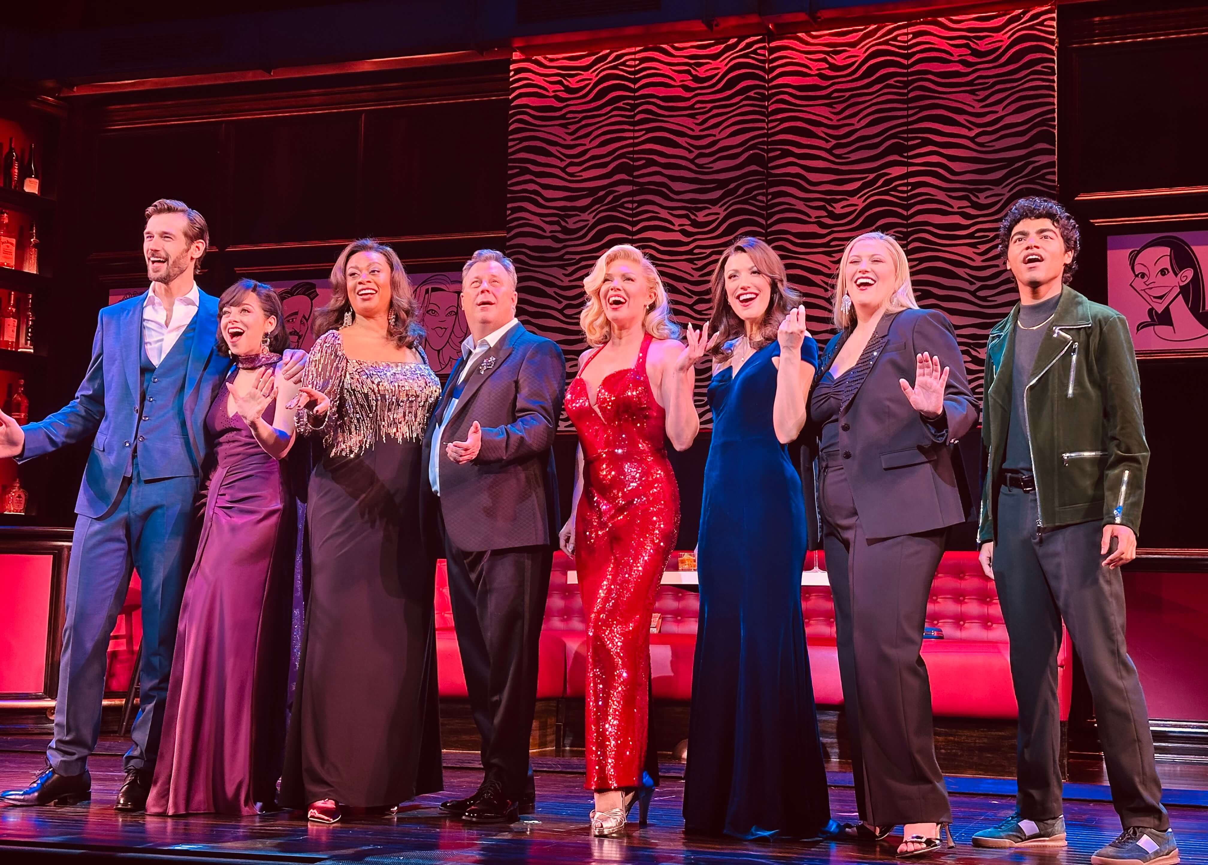 John Behlmann, Krysta Rodriguez, Jacqueline B. Arnold, Brooks Ashmanskas, Robyn Hurder, Caroline Bowman, Bella Coppola, and Nicholas Matos stand together onstage in formal costumes, smiling mid-performance with a bright red and zebra-patterned backdrop behind them.