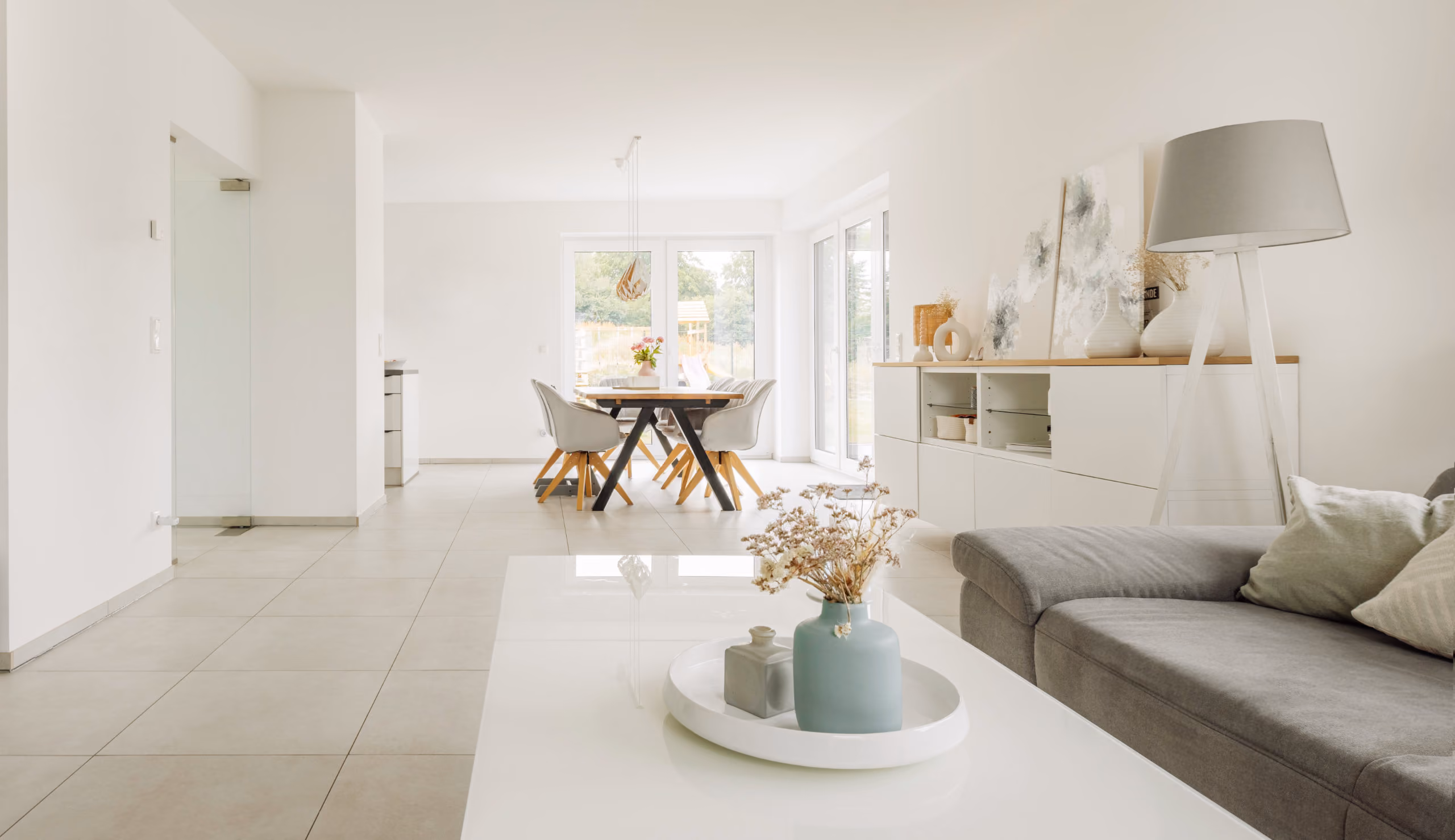 Moderner heller Wohn- und Essbereich mit grauem Sofa, weißem Tisch und großem Fenster.