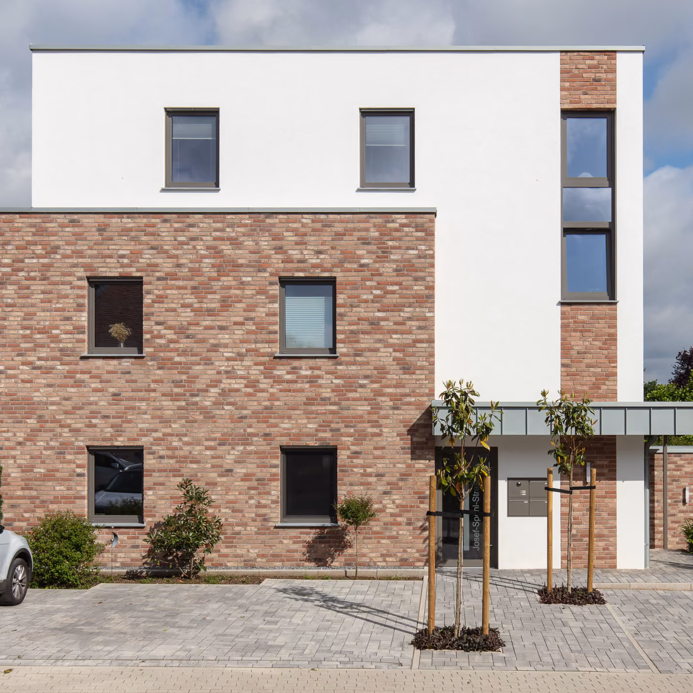 Modernes Wohngebäude mit weißer Fassade, roten Ziegelwänden, Fenster und jungen Bäumen vor dem Haus.