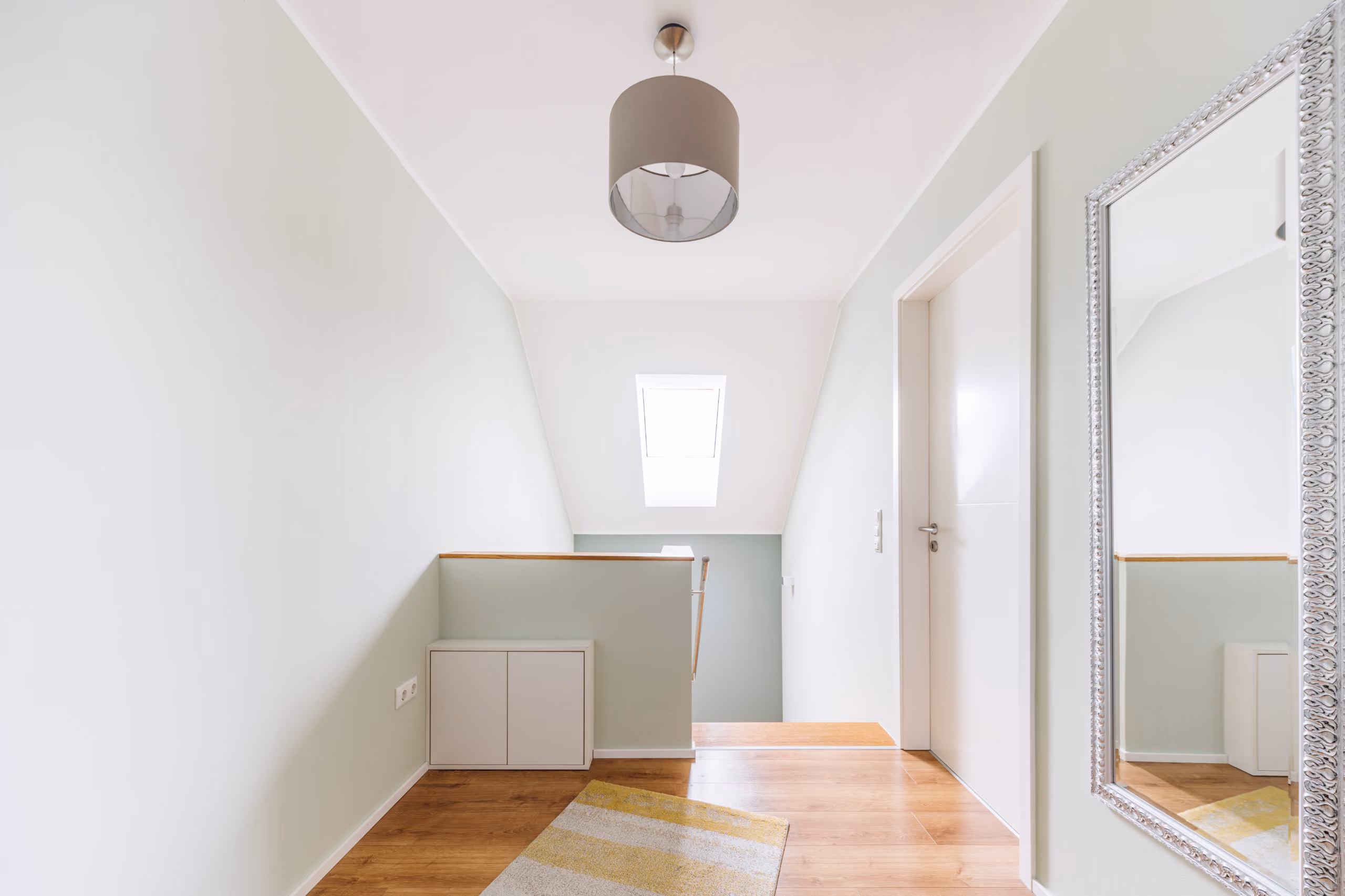 Heller Flur mit Treppe, hellem Holzboden, Teppich, Spiegel und weißer Tür unter Dachfenster.