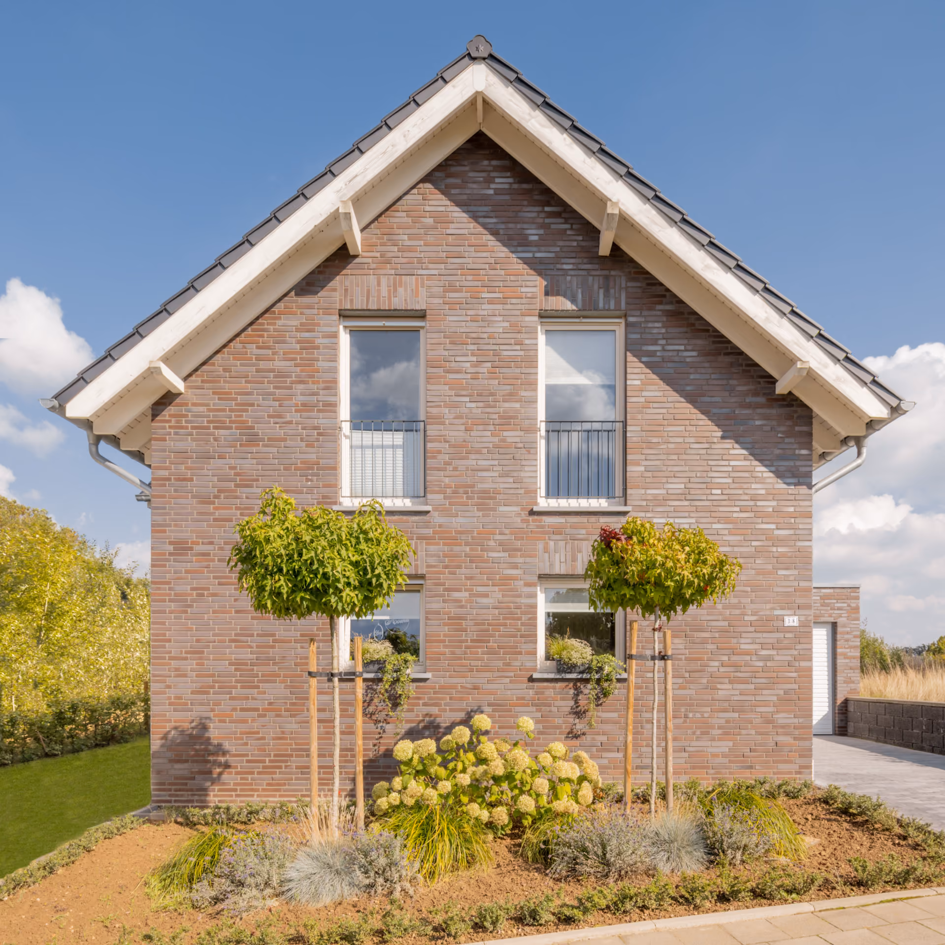 Fassade eines Hauses mit roten Backsteinen, vier Fenstern und gepflegtem Vorgarten mit Blumen und Büschen.