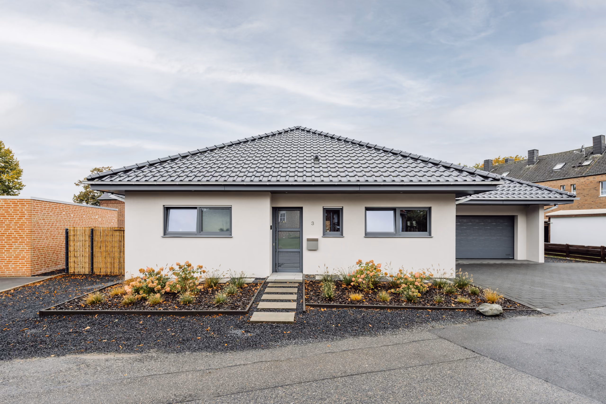 Ein modernes einstöckiges Haus mit grauem Ziegeldach, Pflasterweg und zwei blühenden Beeten.