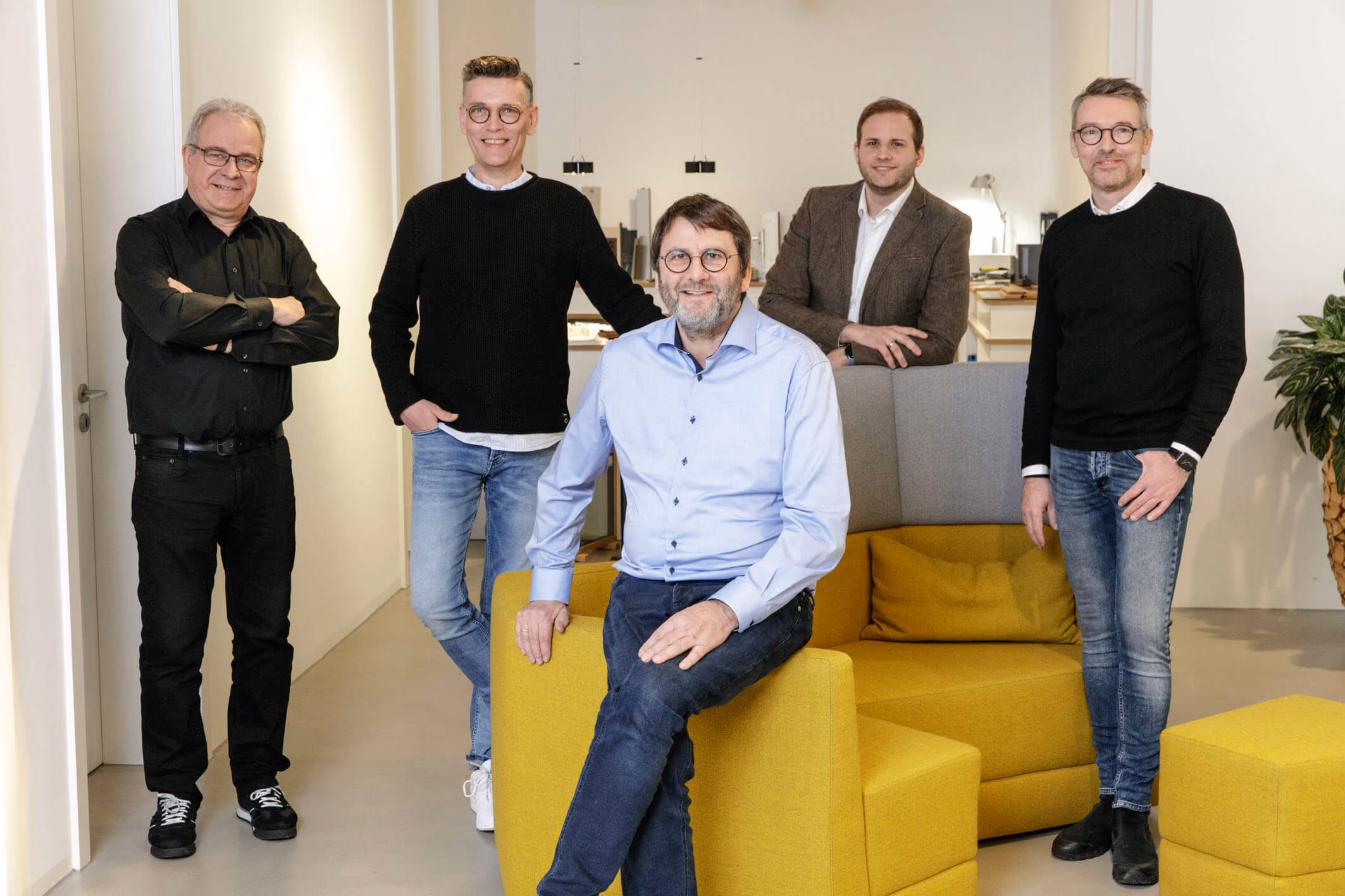 Fünf Männer stehen und sitzen in einem modernen Büro mit gelber Couch und Hockern.