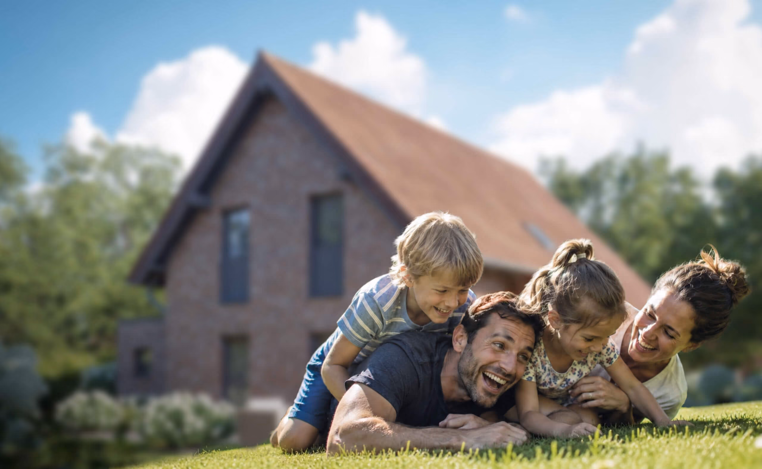 Eine lachende Familie mit zwei Kindern liegt zusammen auf dem Rasen vor einem Haus mit rotem Dach.