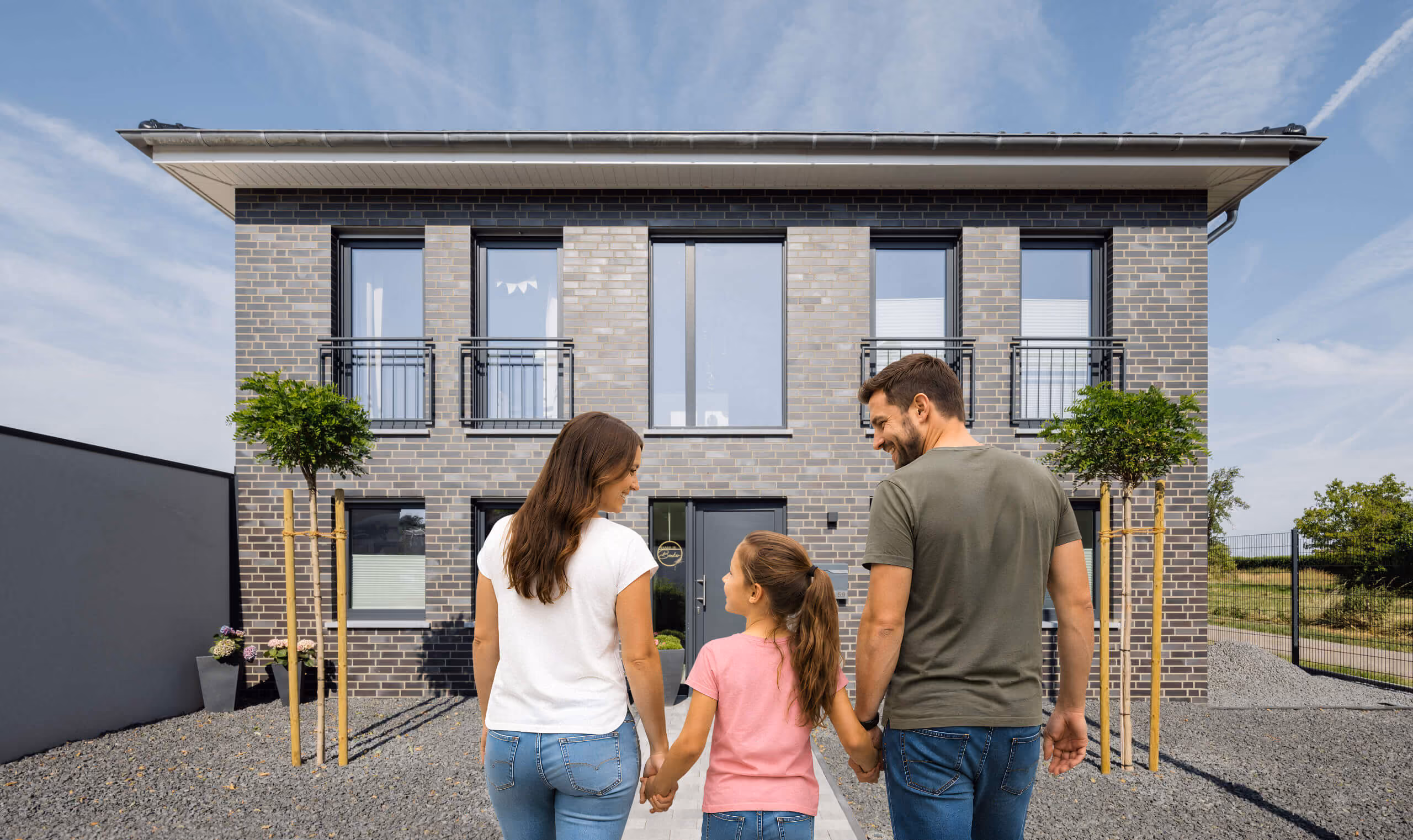 Junge Familie hält Hände vor einem modernen grauen Backsteinhaus mit großen Fenstern.