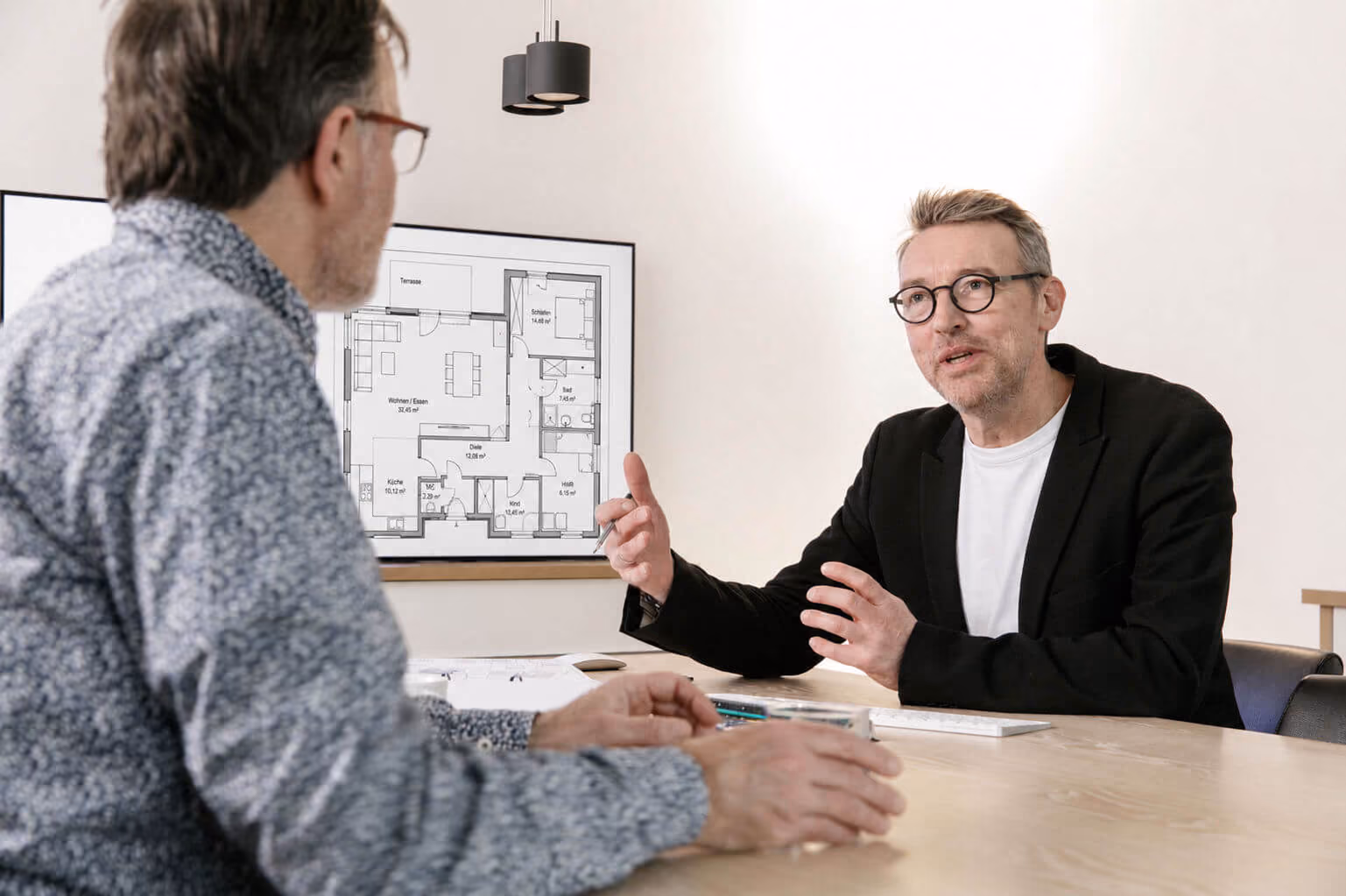 Zwei Männer sprechen an einem Tisch, ein Mann erklärt einen Grundrissplan an der Wand.