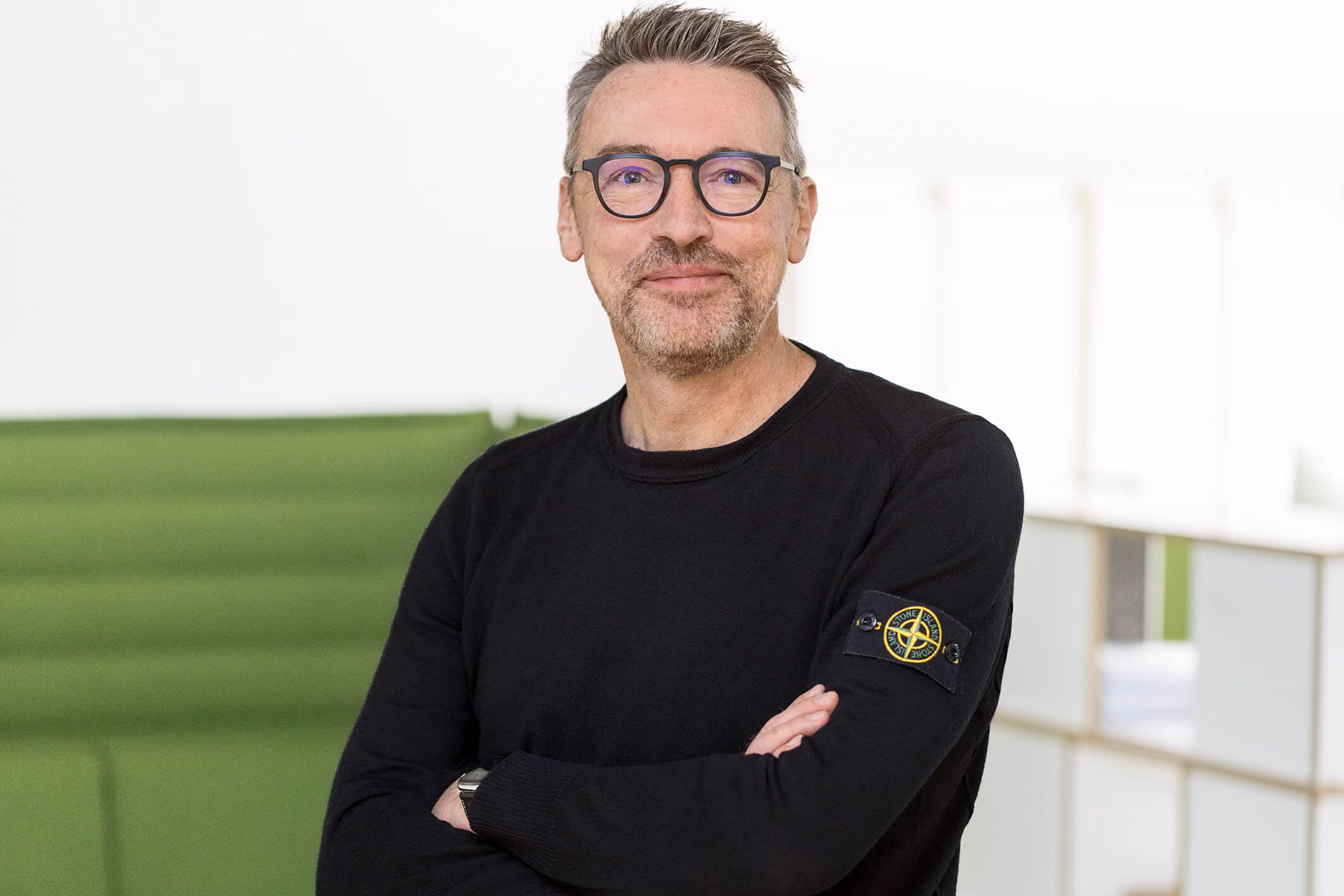 Mann mit Brille und schwarzem Pullover, verschränkte Arme, lächelt vor grünem und weißem Hintergrund.