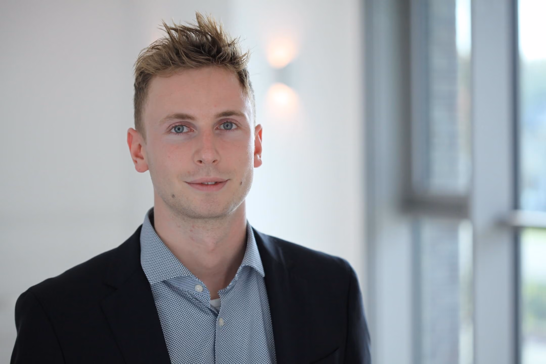 Junger Mann mit blauen Augen und zerzaustem Haar in blauem Hemd und schwarzem Sakko.