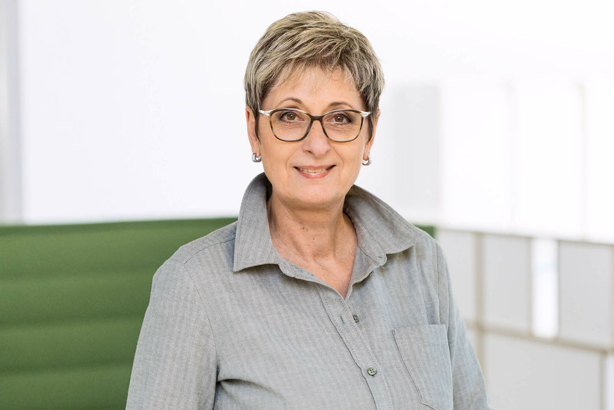 Ältere Frau mit Brille und grauen Haaren lächelt, trägt Hemd mit offenem Kragen.