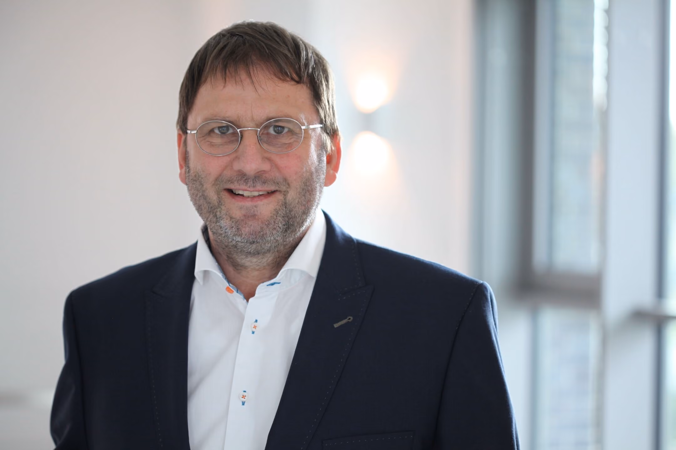 Mann mit kurzer brauner Haare, Brille und Bart, trägt weißen Hemd und blauen Anzug.