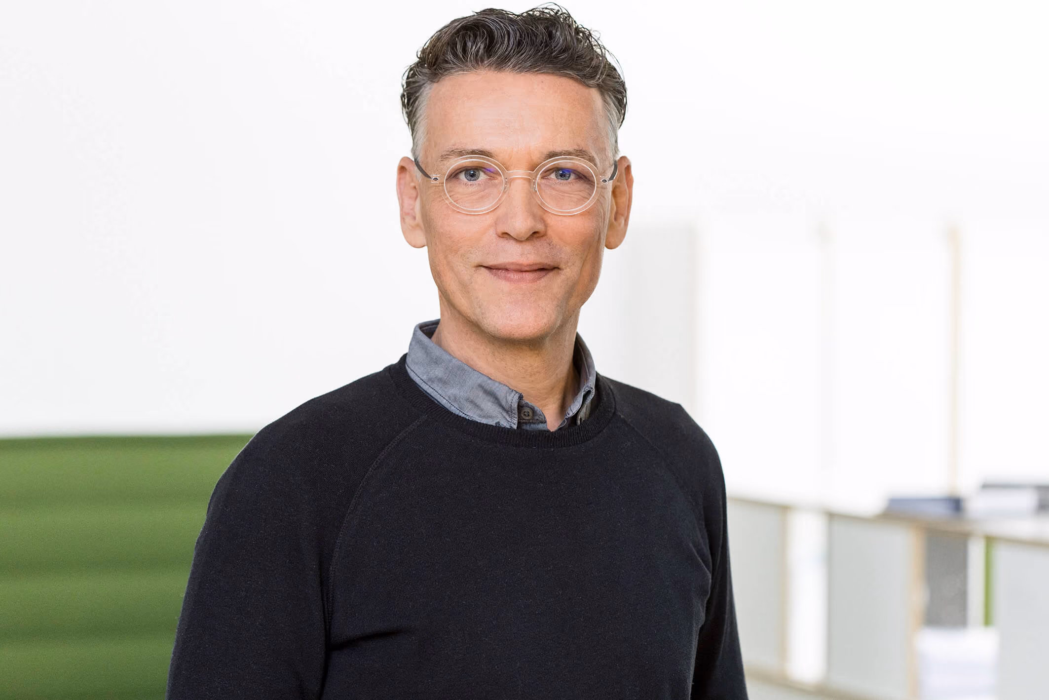 Porträt eines Mannes mit Brille, grauen Haaren, blauem Hemd und schwarzem Pullover vor grünem Hintergrund.