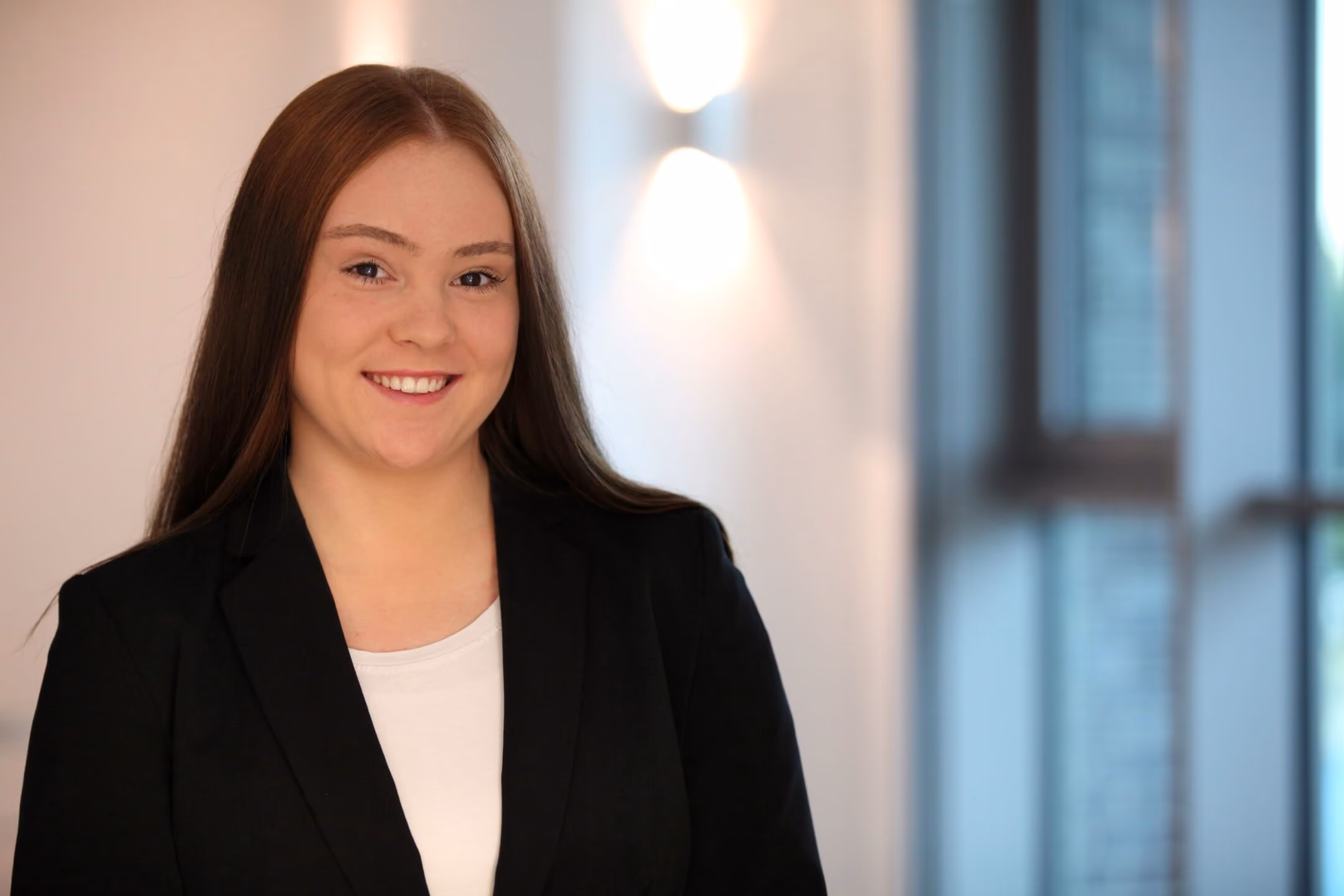 Junge Frau mit langem braunem Haar trägt schwarzen Blazer und weiße Bluse, lächelnd vor einem neutralen Hintergrund.
