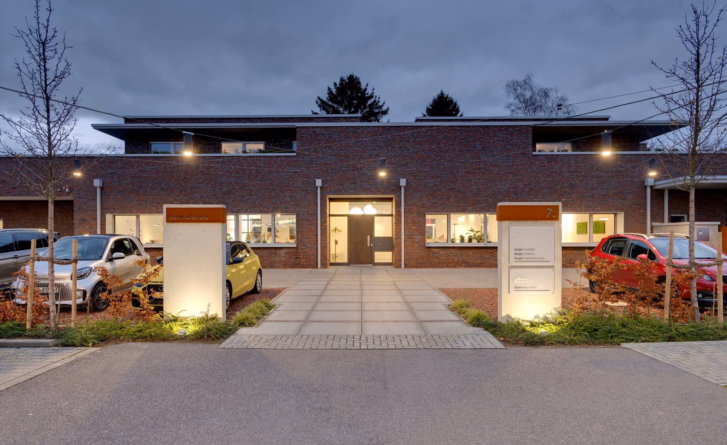 Beleuchteter Eingang eines modernen Backsteingebäudes mit geparkten Autos links und rechts.
