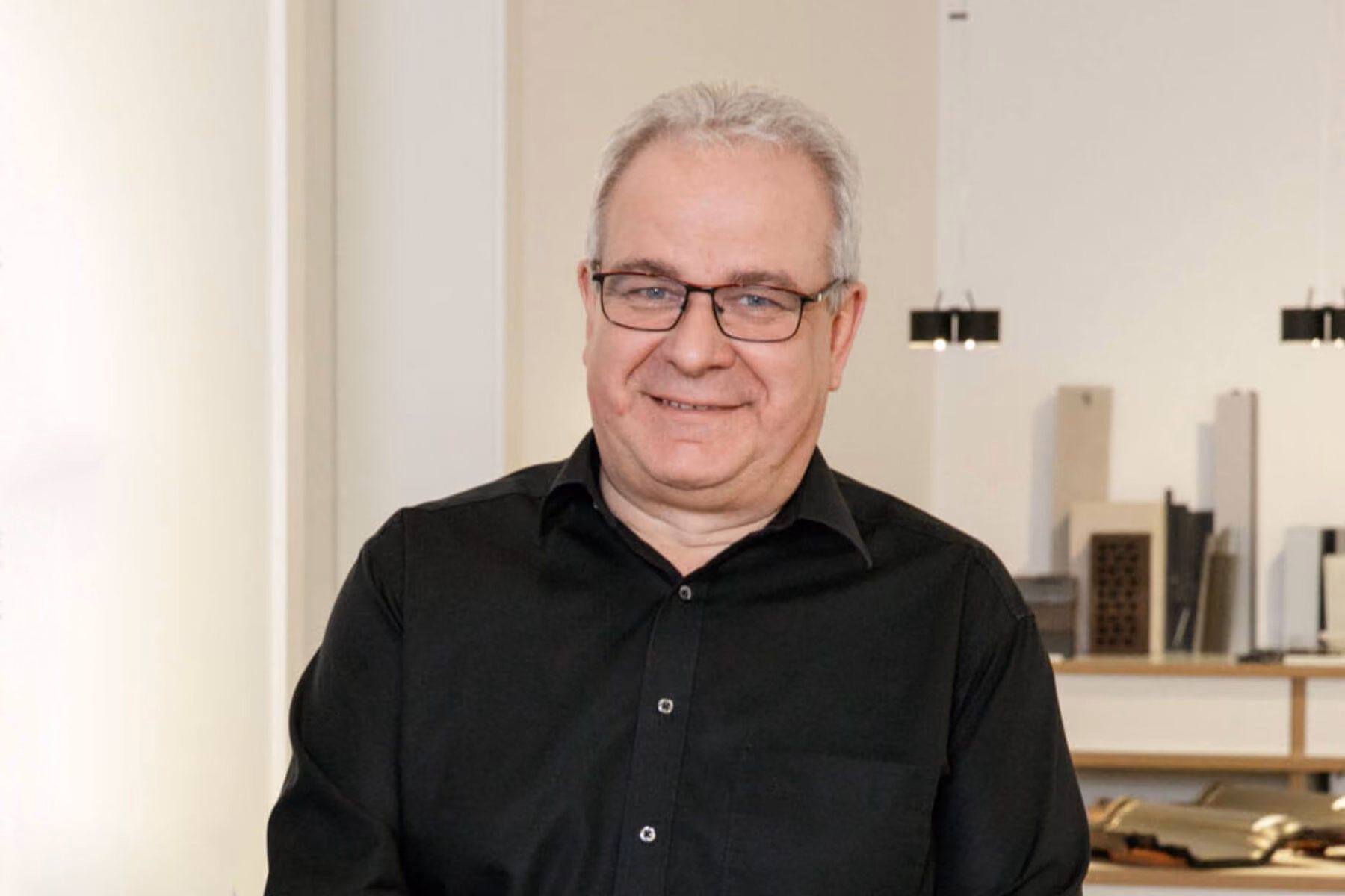 Ein älterer Mann mit grauem Haar und Brille trägt ein schwarzes Hemd und lächelt.