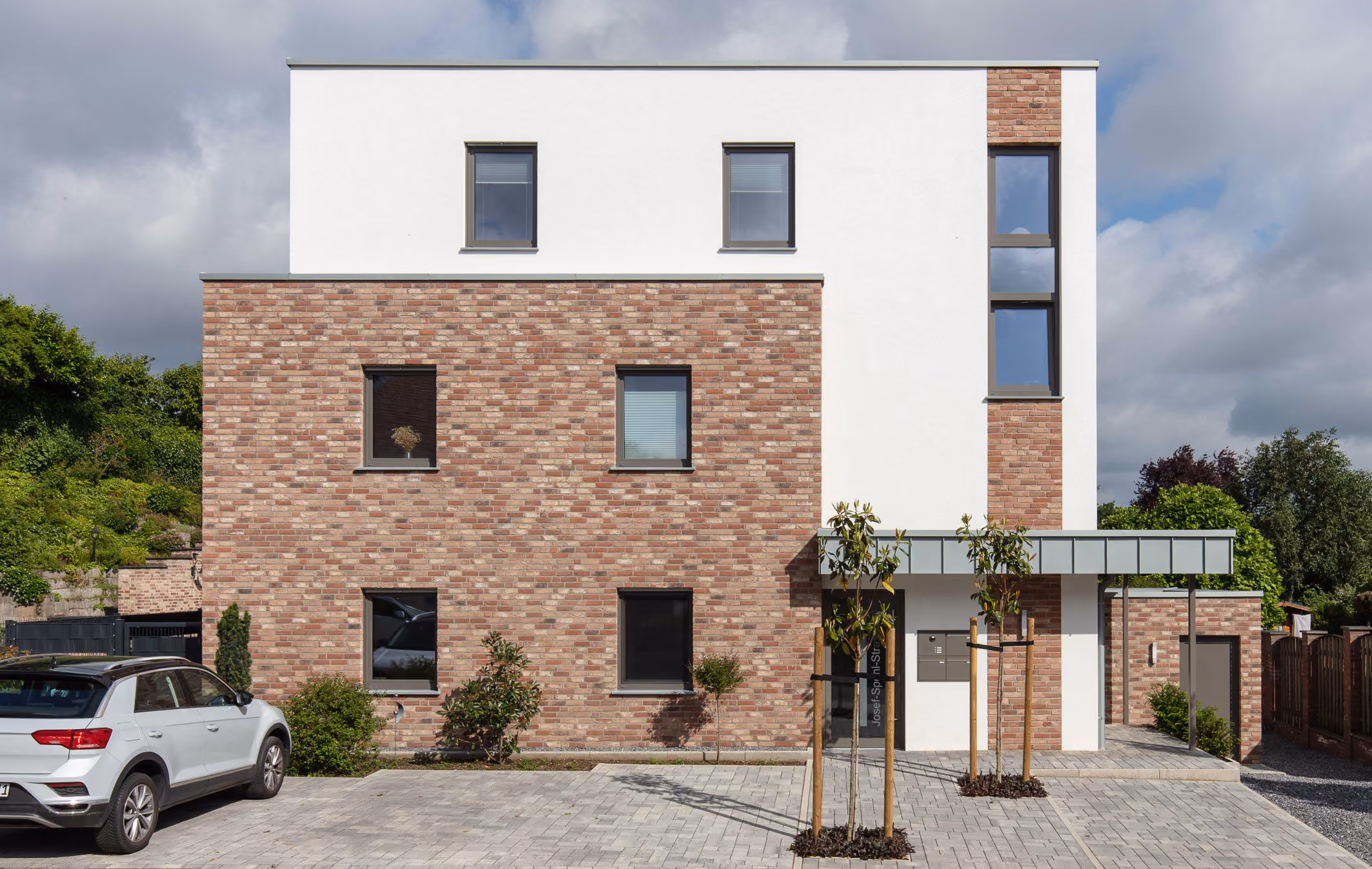 Modernes kubisches, mehrstöckiges Wohngebäude mit weißer und ziegelroter Fassade, Autos und Bäumen davor.