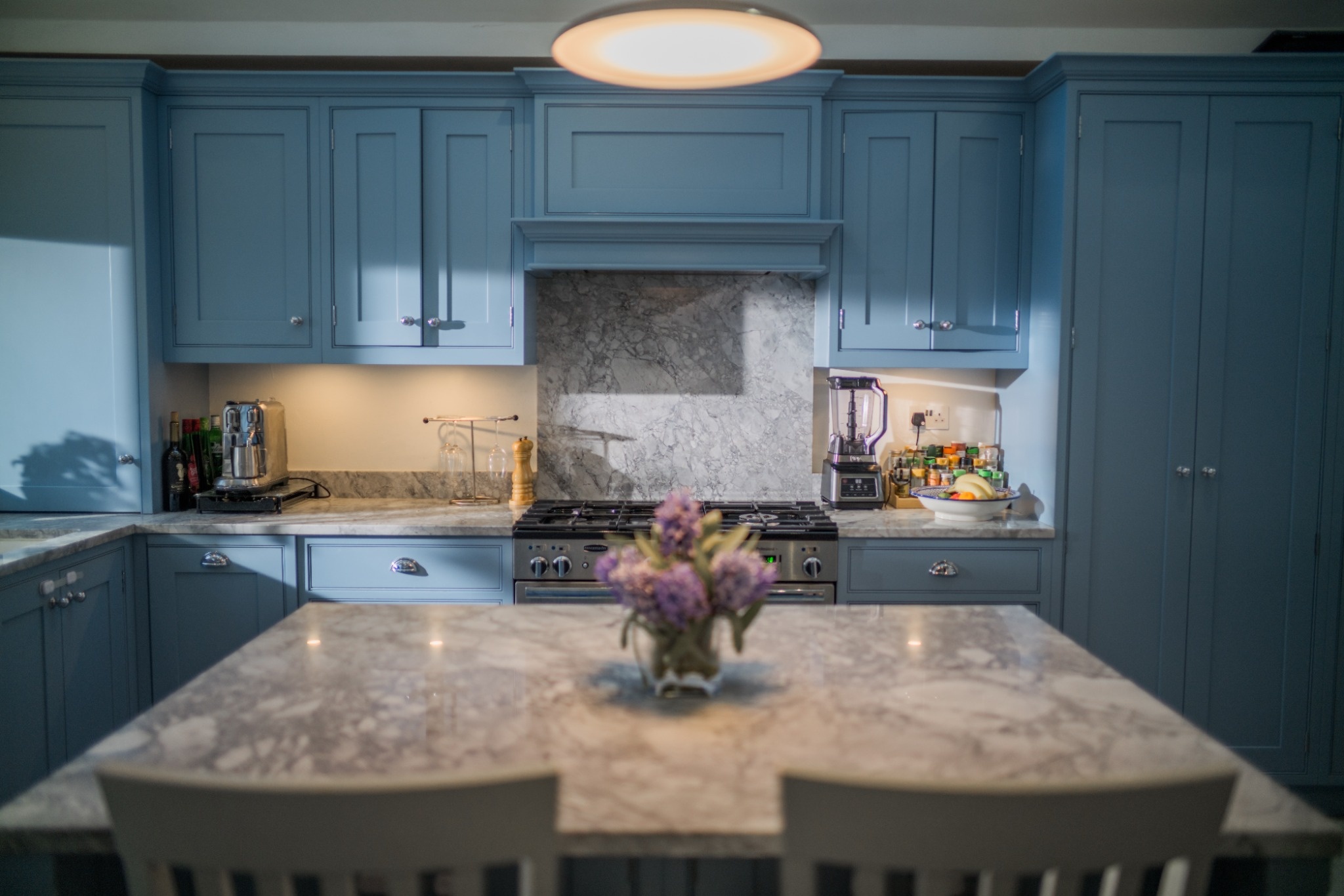 Modern kitchen with blue cabinets, marble countertops, stovetop, blender, and a flower vase on island.