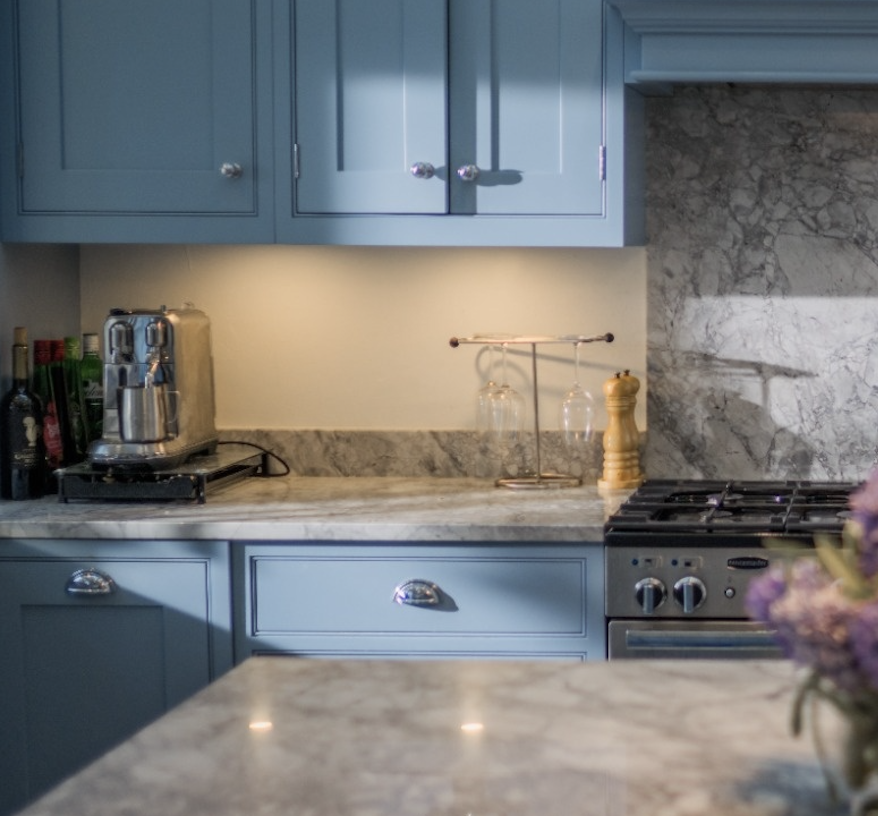 Marble countertop kitchen with blue cabinets, espresso machine, wine bottles, and gas stove.