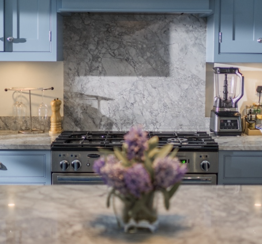 Kitchen with marble countertops, blue cabinets, gas stove, blender, and blurred purple flowers in foreground.