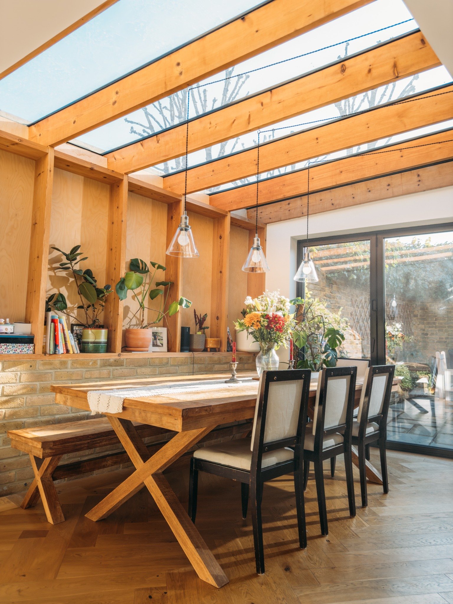 Dining area with wooden table, bench, four chairs, hanging lights, plants, and large glass doors.