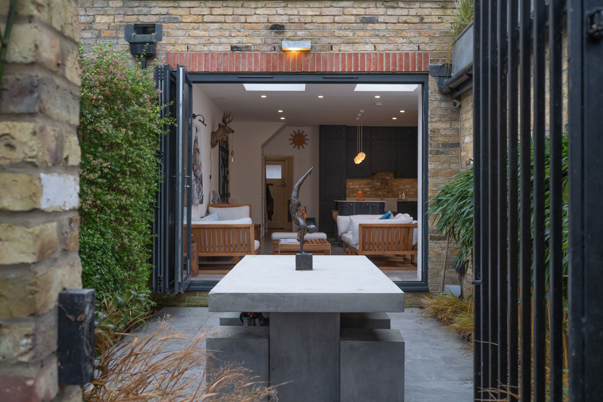 Concrete outdoor table and benches with a sculpture, leading to an open living room with wooden furniture.