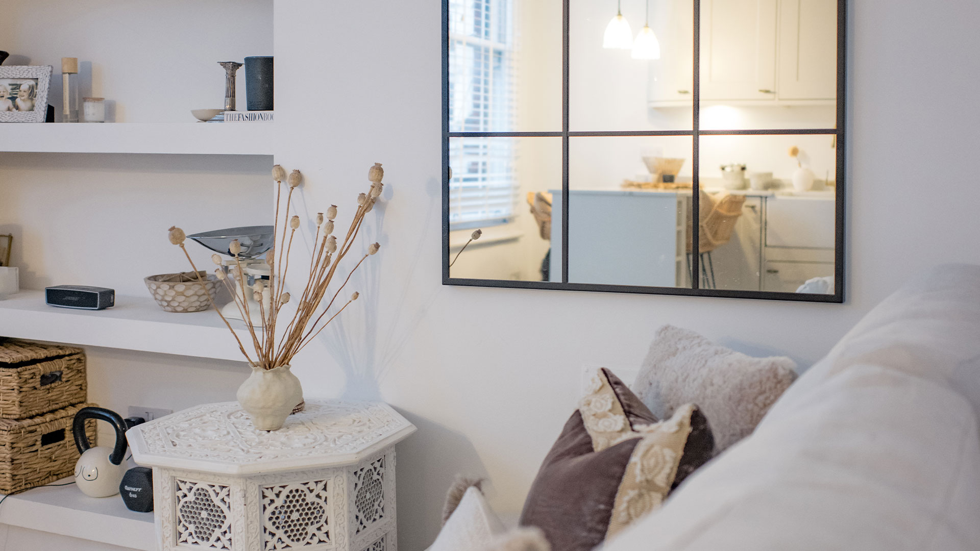 Cozy living room corner with a carved white table, dried plants, pillows, and a window-like mirror.