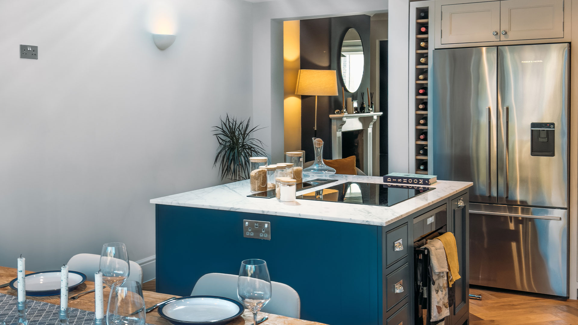 Modern kitchen with blue island, marble countertop, stainless steel fridge, and dining table set with plates and glasses.