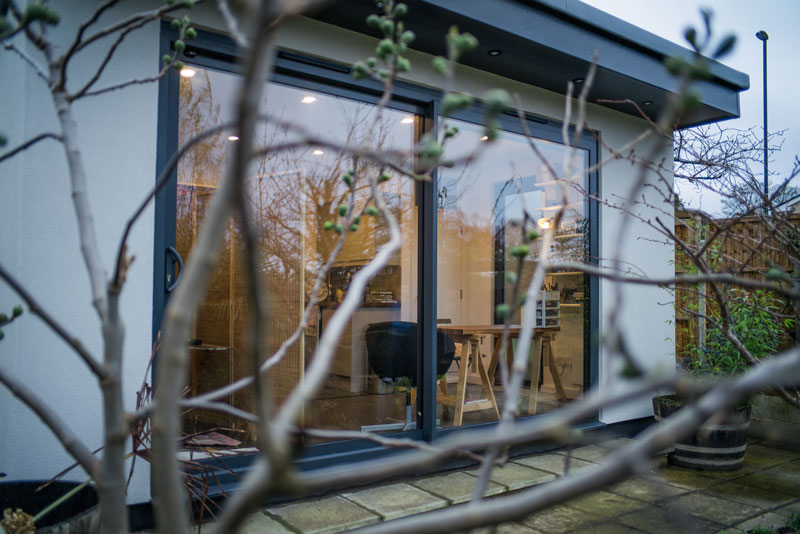 View through tree branches of a modern room with large sliding glass doors at dusk.