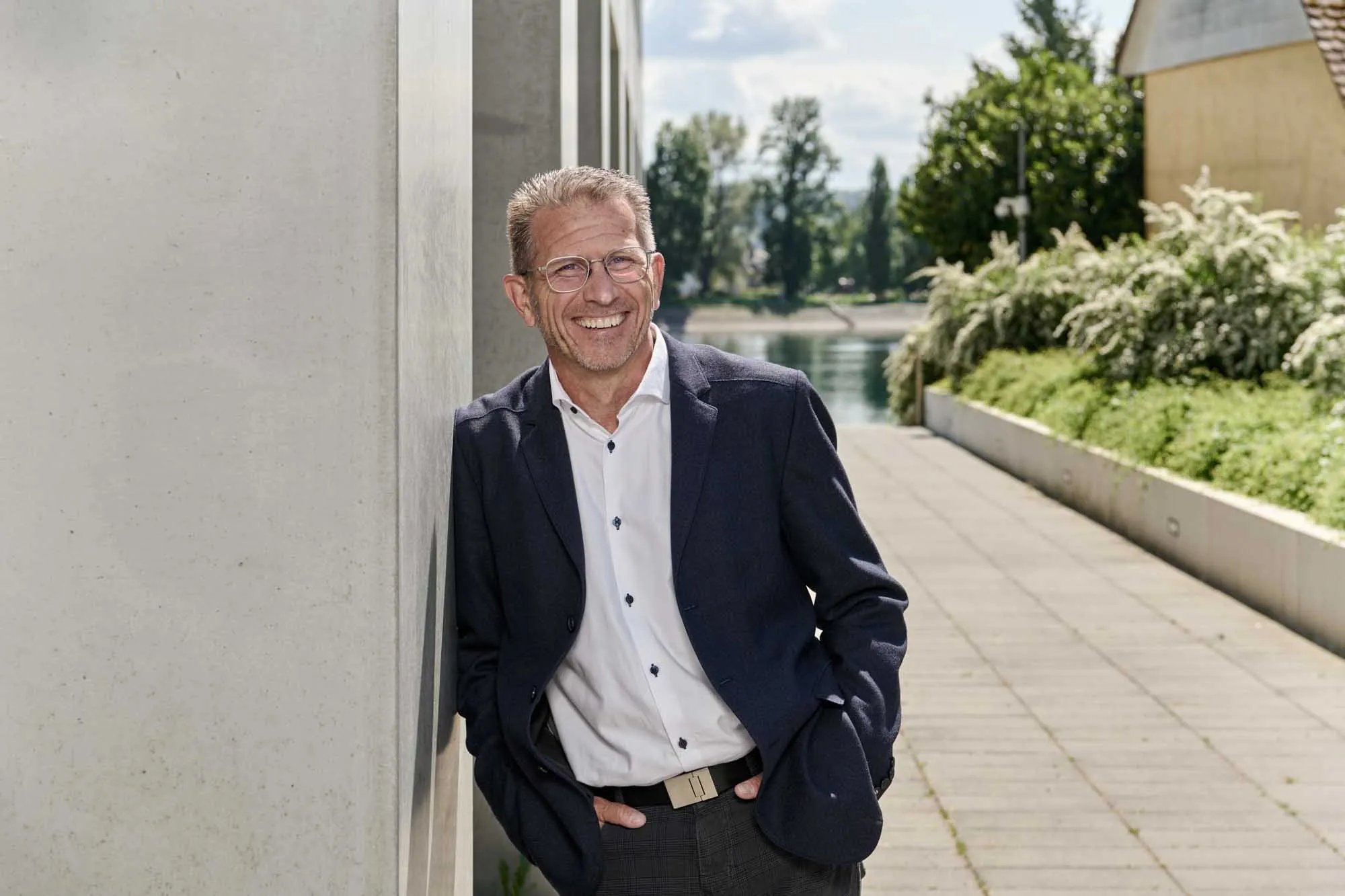 Lächelnder Mann mit Brille lehnt entspannt an einer Wand im Freien, mit Wasser und Bäumen im Hintergrund.