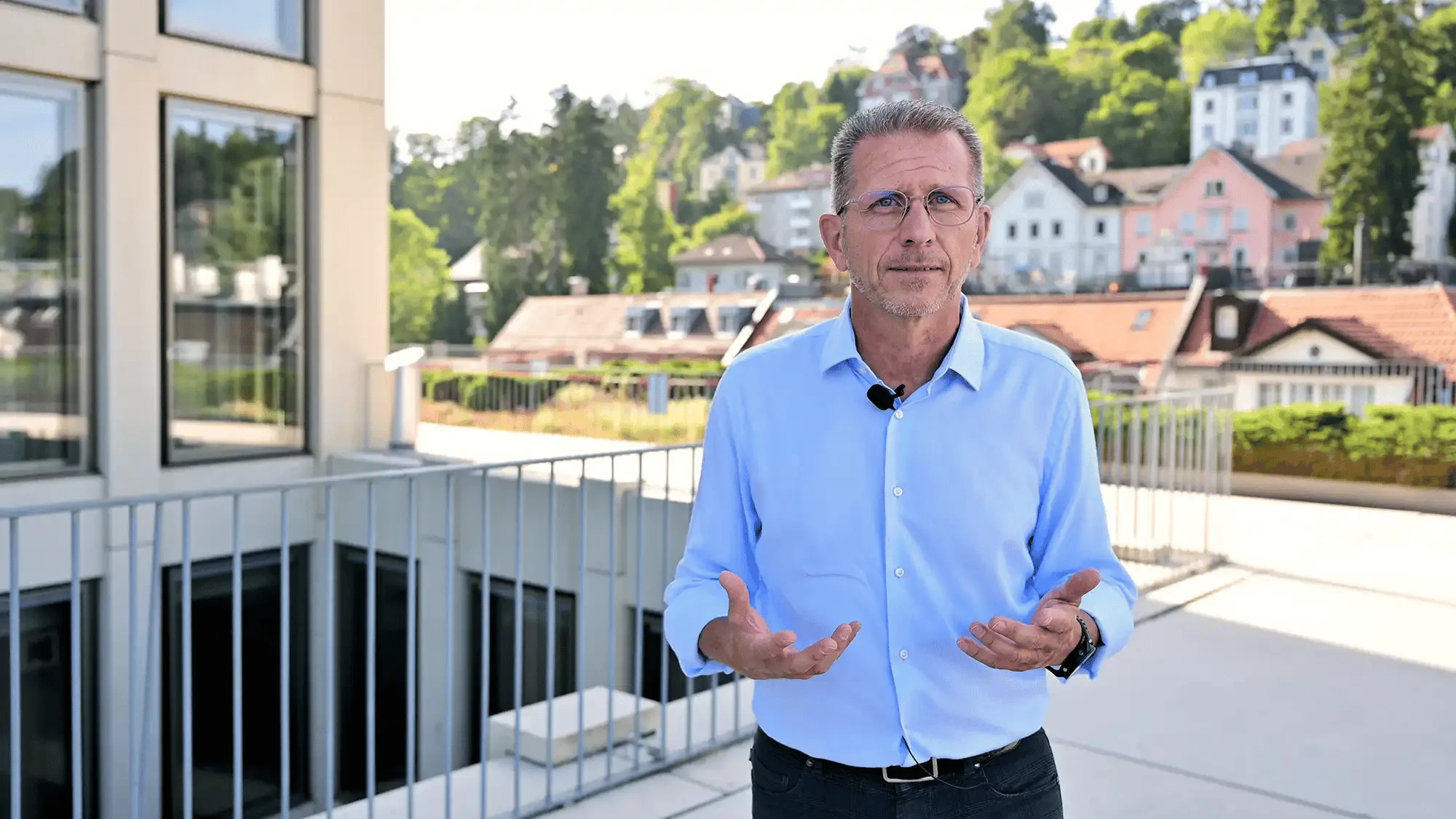 Mann in blauem Hemd mit Brille spricht draußen auf Balkon mit Haus- und Baumblick im Hintergrund.