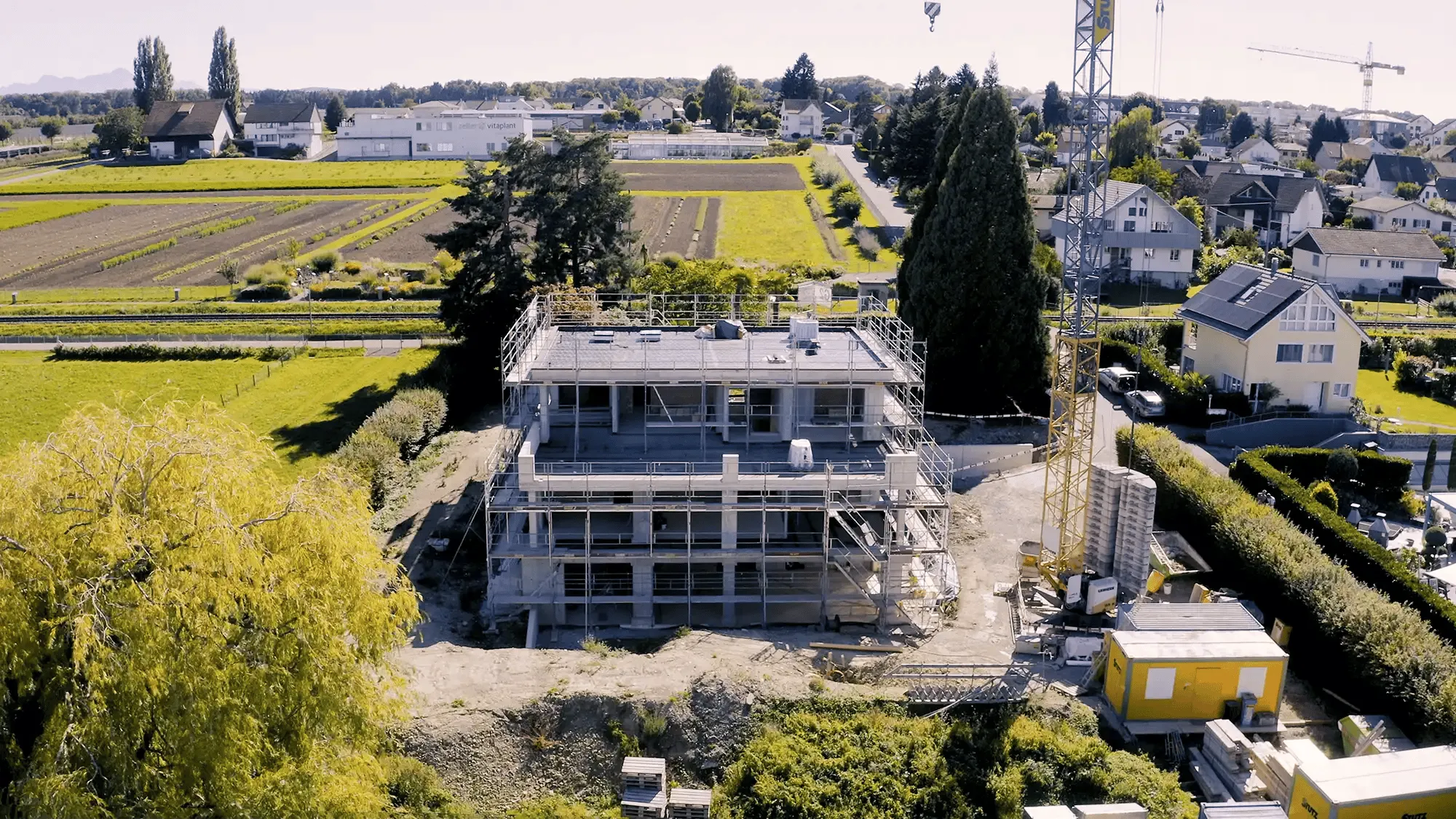 Mehrstöckiges Rohbaugebäude mit Gerüst und Baukran in einem ruhigen Vorort mit ländlichem Hintergrund.