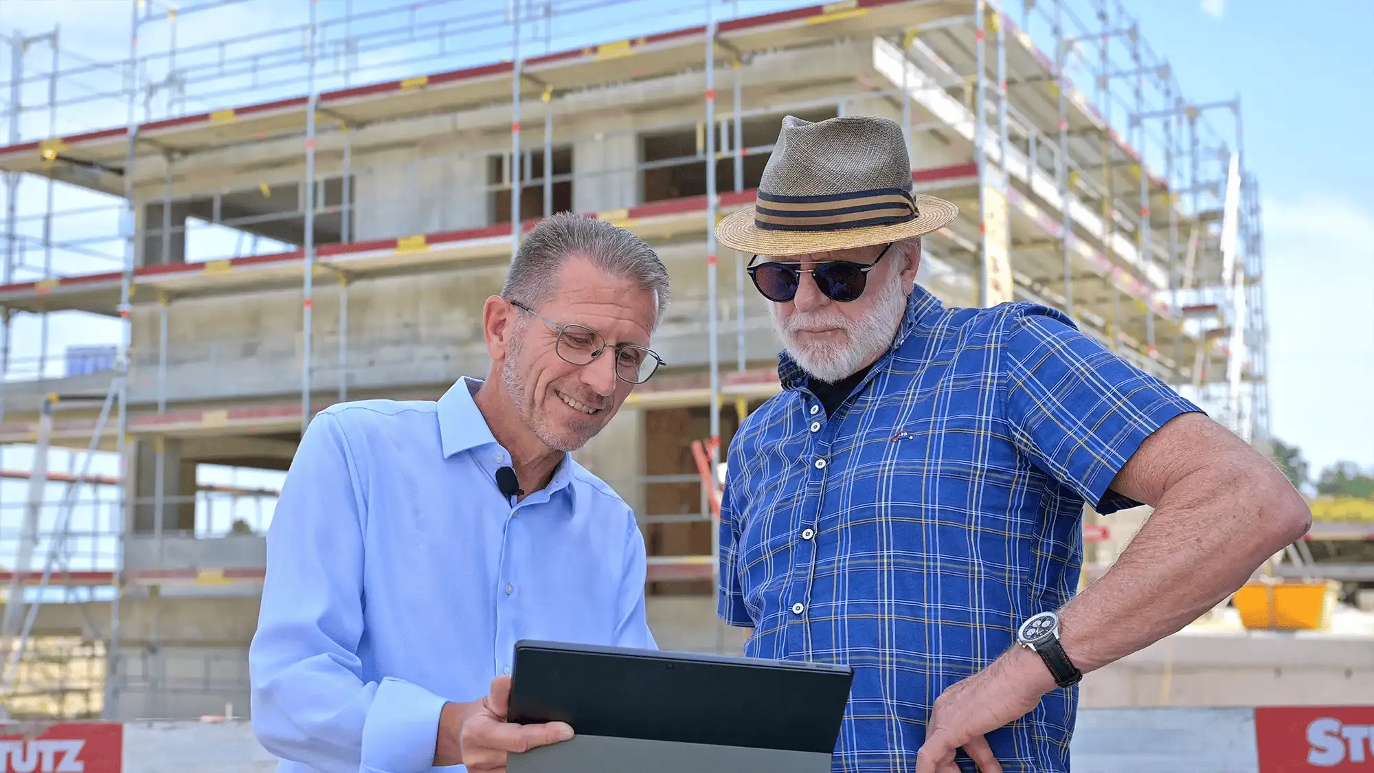 Zwei Männer betrachten Tablet vor einem Rohbau mit Gerüst, einer trägt Hut und Sonnenbrille.