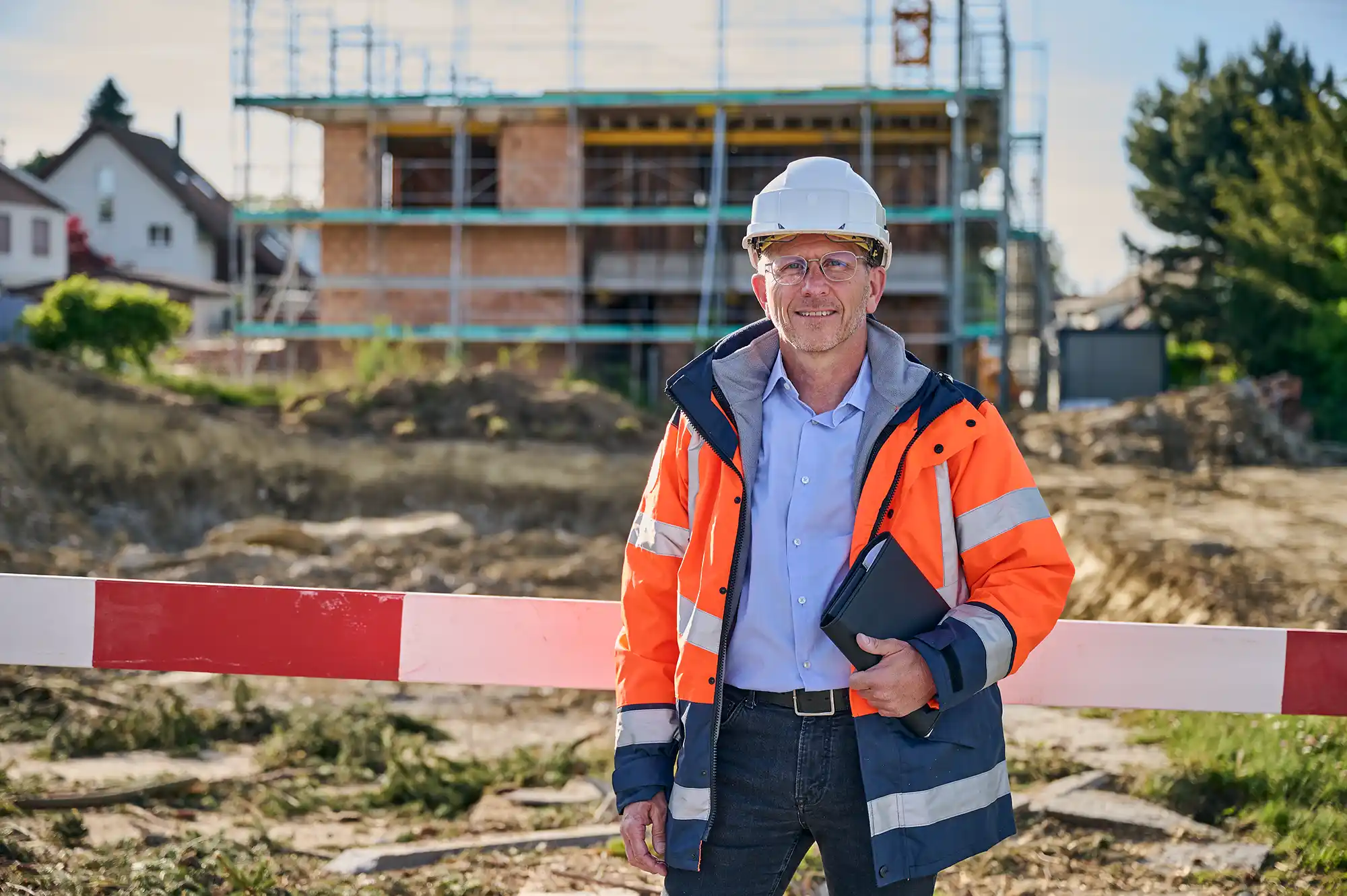 Benjamin Wulf: Bauleiter mit Schutzhelm und Warnjacke hält Klemmbrett auf einer Baustelle mit unfertigem Gebäude im Hintergrund.