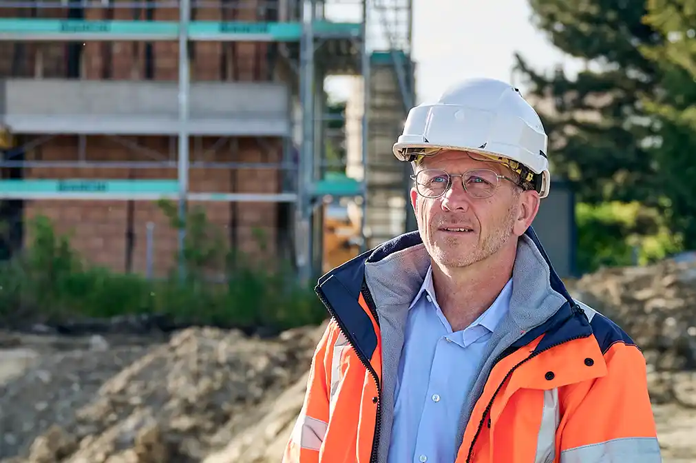 Benjamin Wulf: Bauleiter mit Schutzhelm und Warnjacke hält Klemmbrett auf einer Baustelle mit unfertigem Gebäude im Hintergrund.