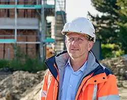 Bauleiter mit Schutzhelm und oranger Sicherheitsjacke auf einer Baustelle im Freien.