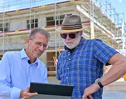 Zwei Männer prüfen gemeinsam eine Bauplanung vor einer im Bau befindlichen Baustelle.