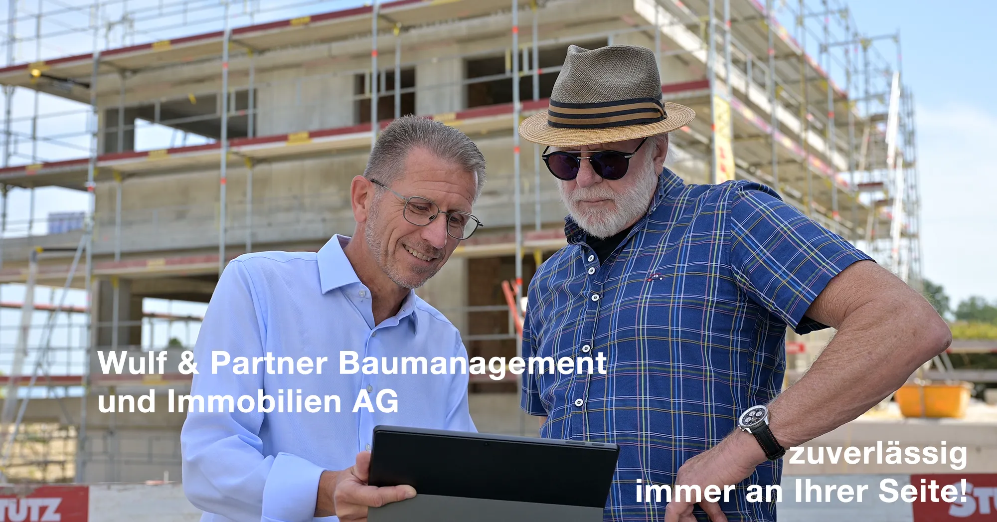Zwei Männer am Bau schauen auf ein Tablet vor einem Rohbau mit Gerüst an einem sonnigen Tag.