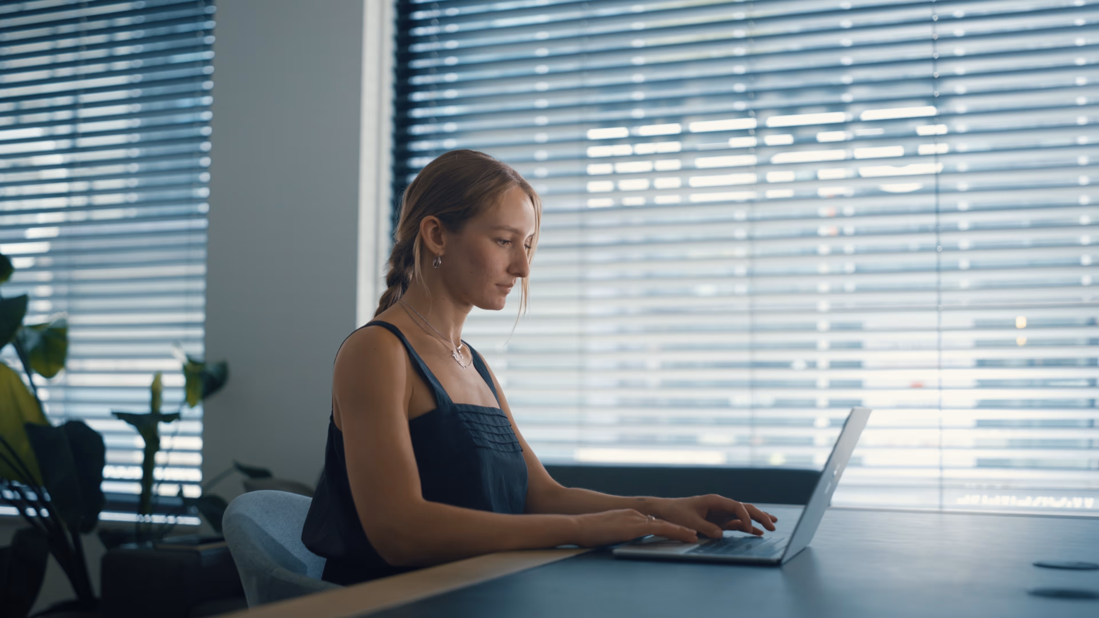 Vrouw die op laptop werkt zittend aan een bureau vóór een raam met jaloezieën.