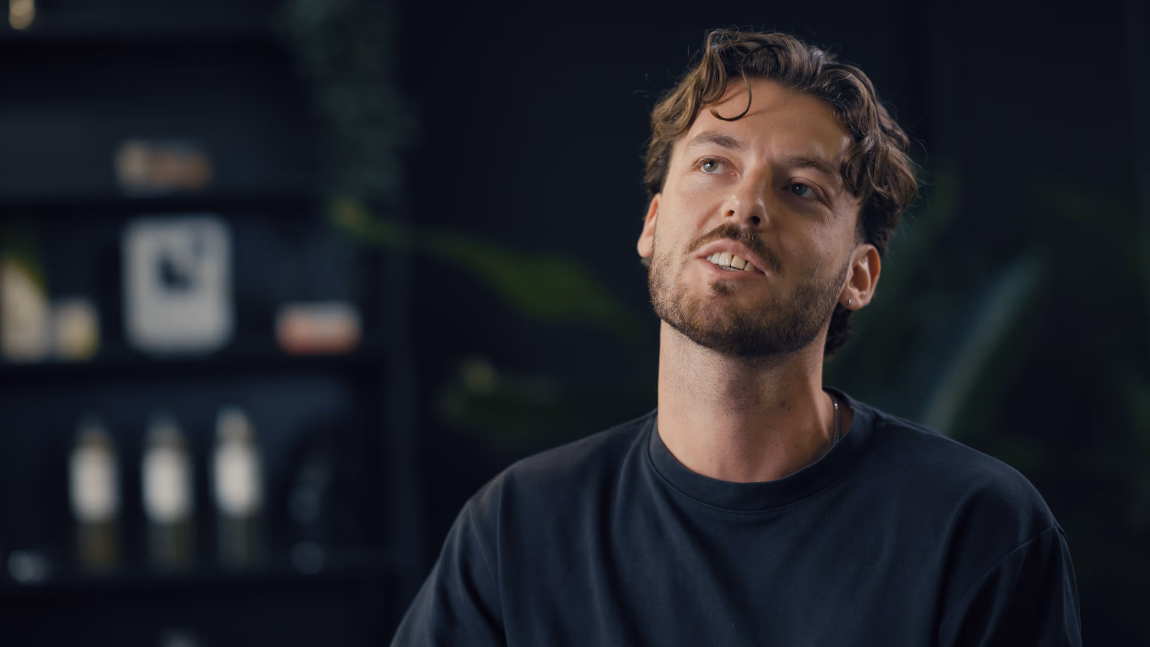 Man met baard en krullend haar kijkt omhoog, draagt een donker shirt in een donkere kamer.