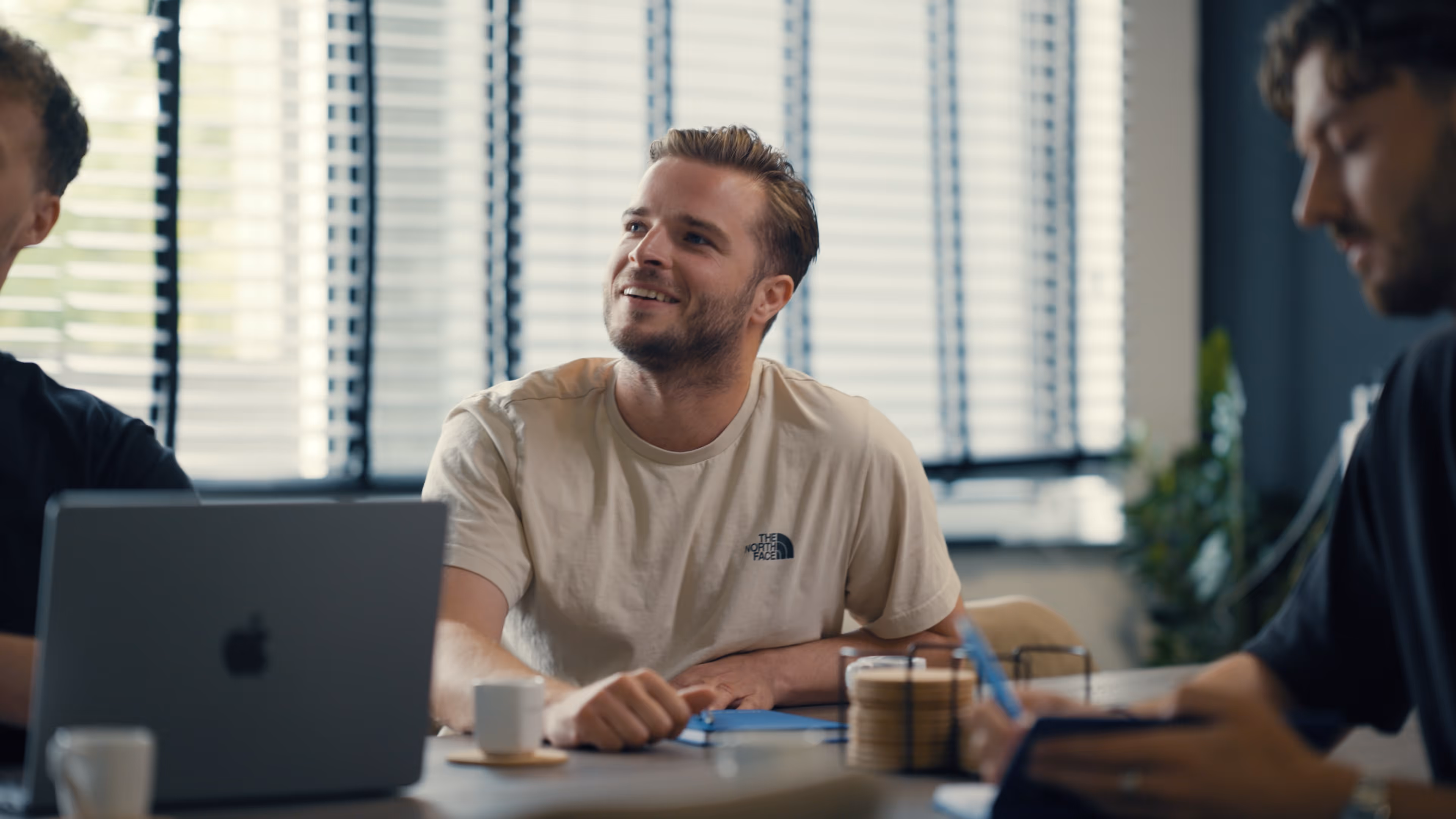 Jonge man die glimlacht tijdens een vergadering met laptop en koffiekopjes op tafel.