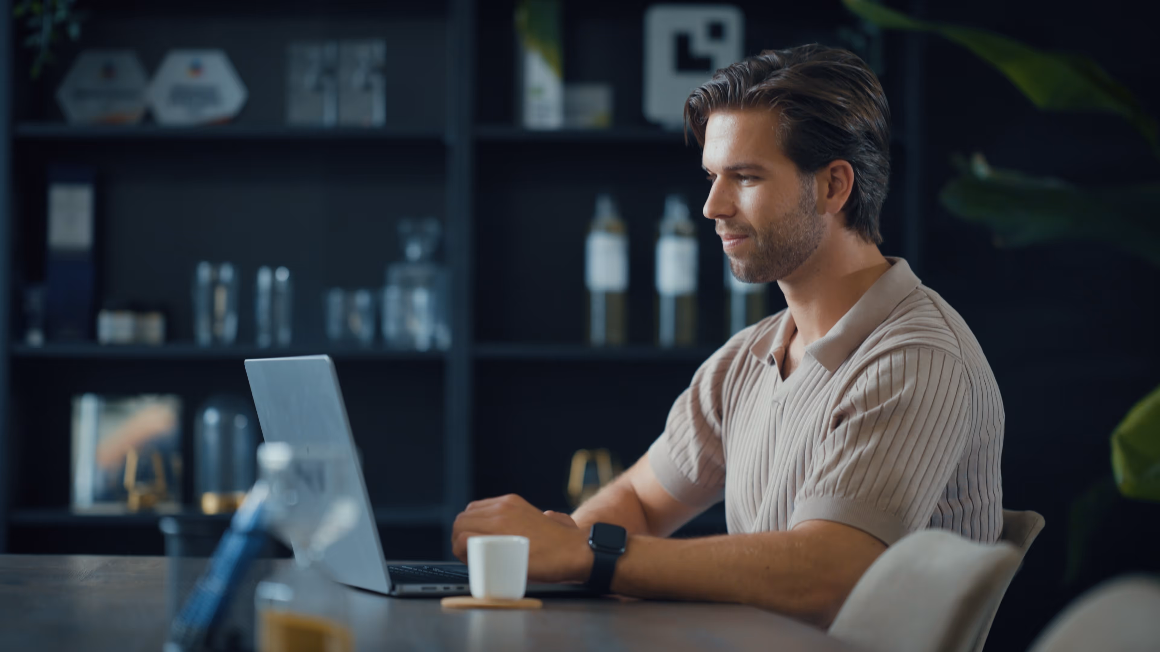 Man in beige shirt working on a laptop at a table with a cup and blurred background.