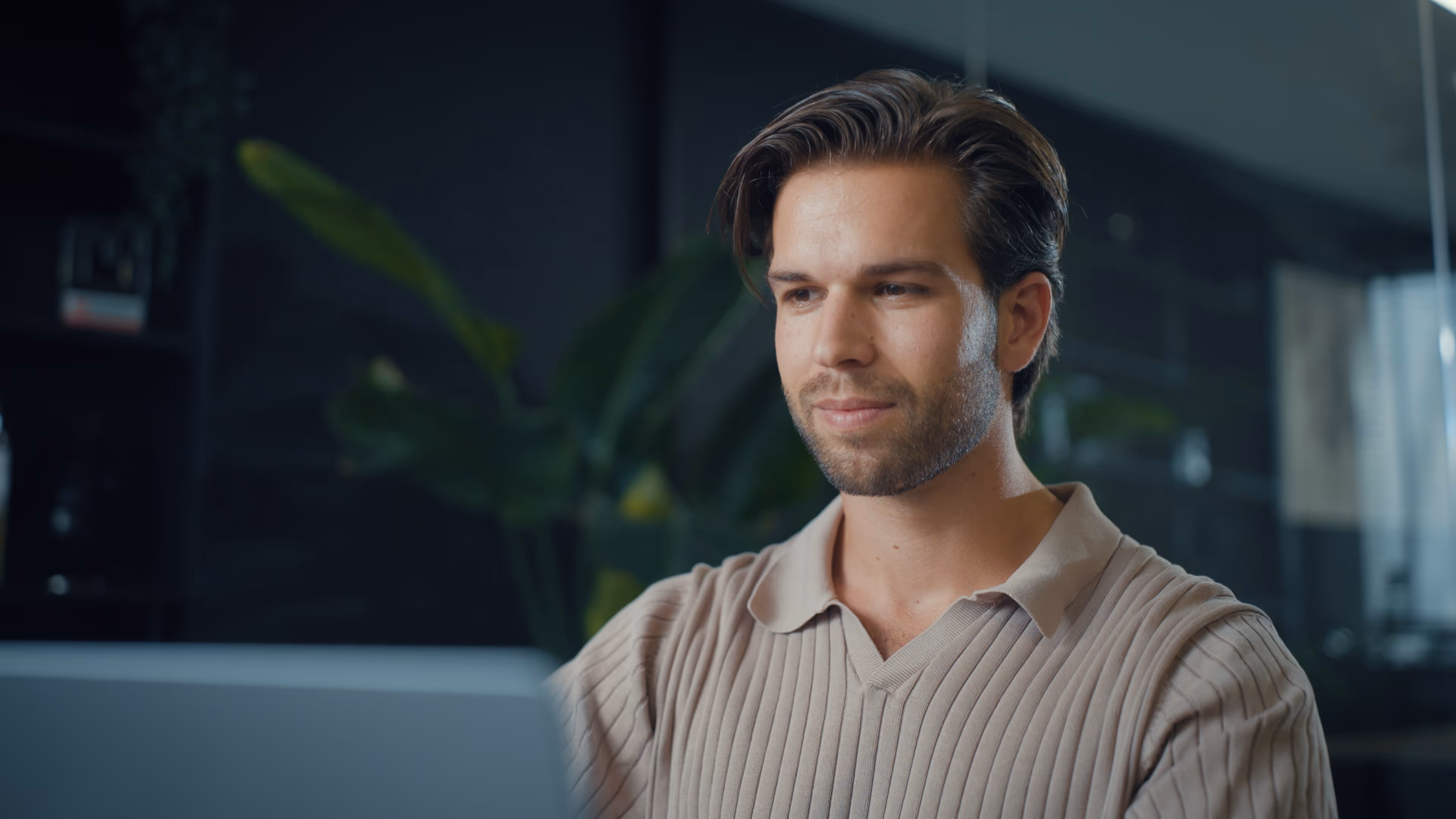Jonge man met beige trui werkt geconcentreerd op laptop in moderne, donkere werkomgeving.