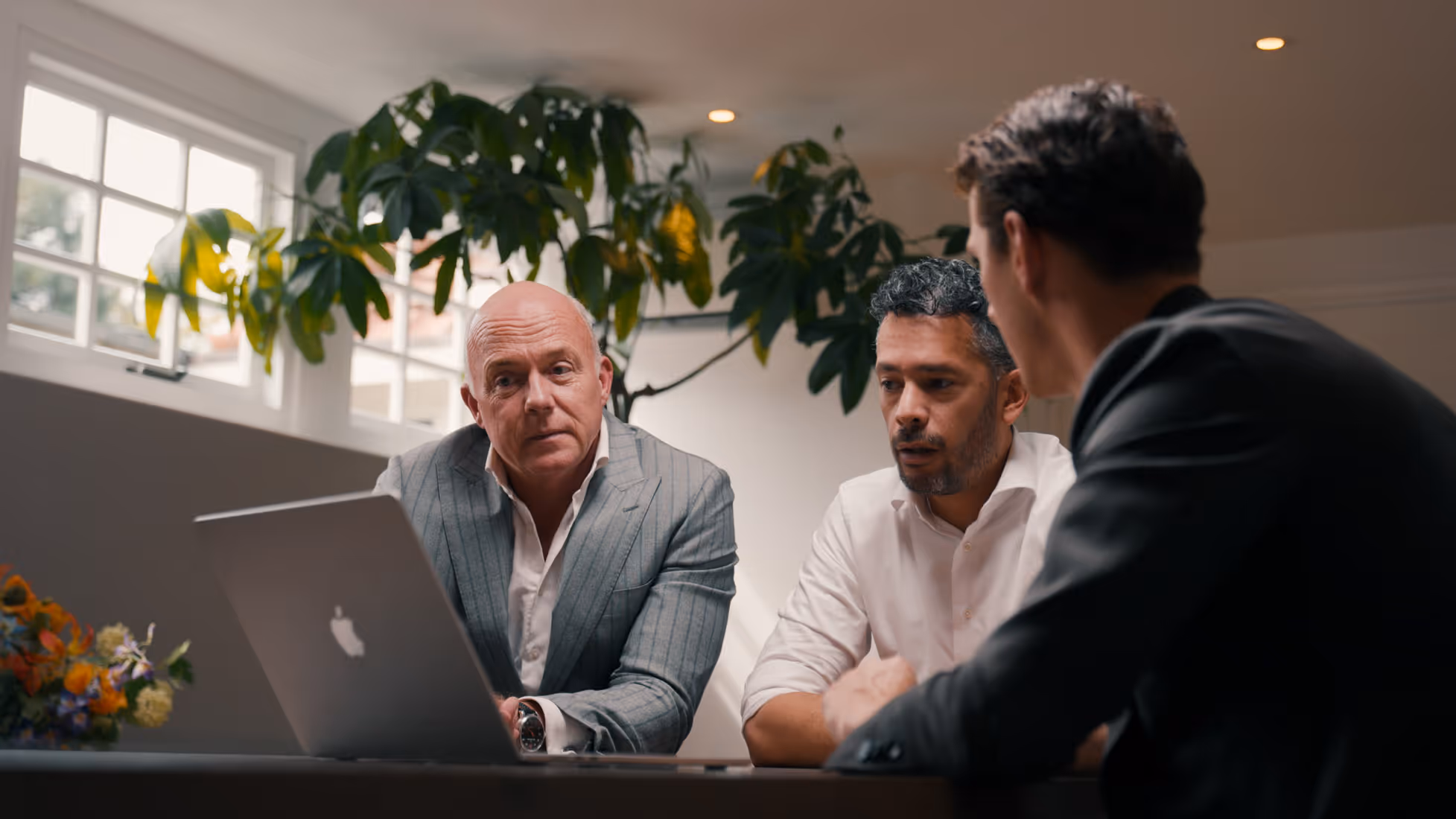 Drie mannen in een zakelijke vergadering die naar een laptop kijken in een lichte kamer.