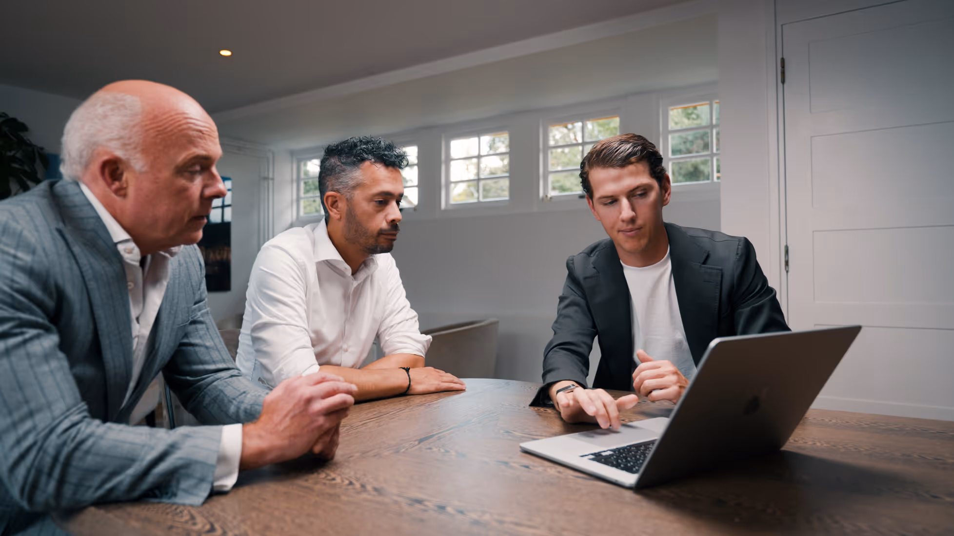 Drie mannen in een bespreking kijken gezamenlijk naar een laptop aan een tafeltje in een kantoor.