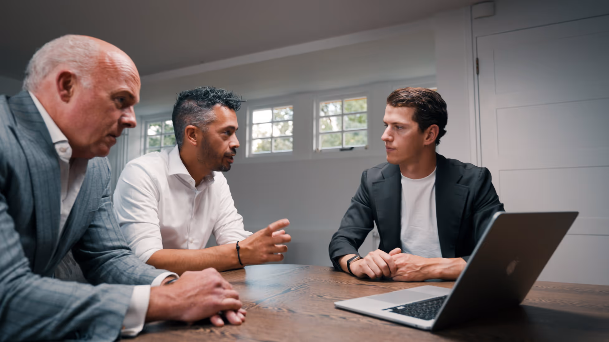 Drie mannen in een vergadering aan een houten tafel met een laptop, verdiept in gesprek.